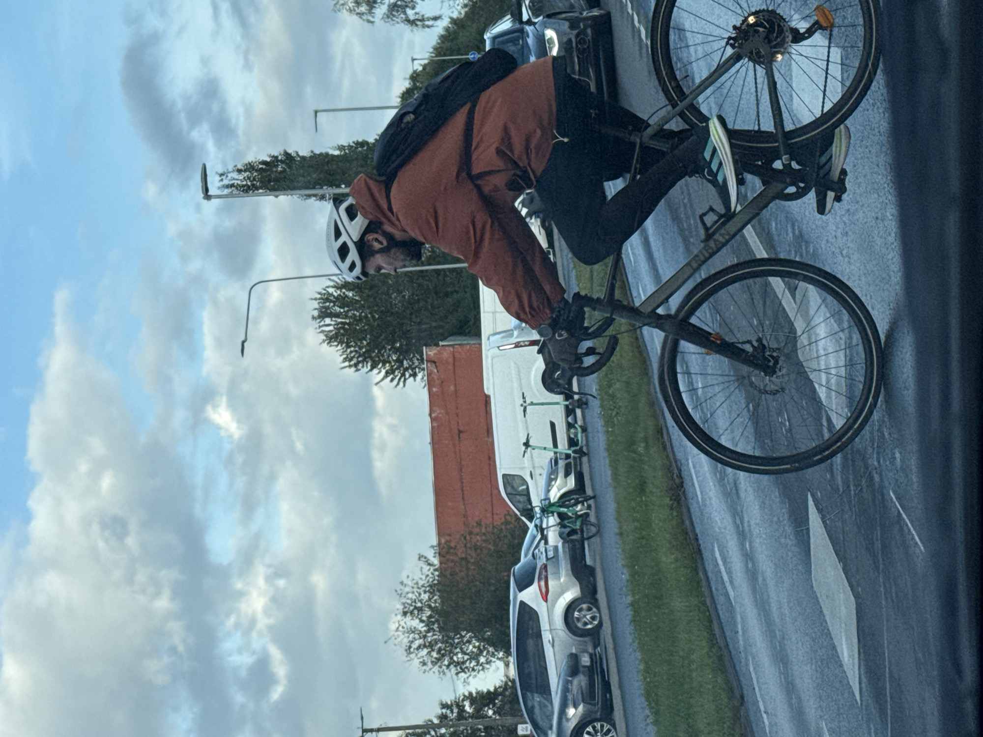 A man wearing a white helmet and an orange jacket rides a bicycle on a wet road in Tallinn, Estonia. He has a black backpack on. In the background, there are cars, vans, and electric scooters. The sky is cloudy.
