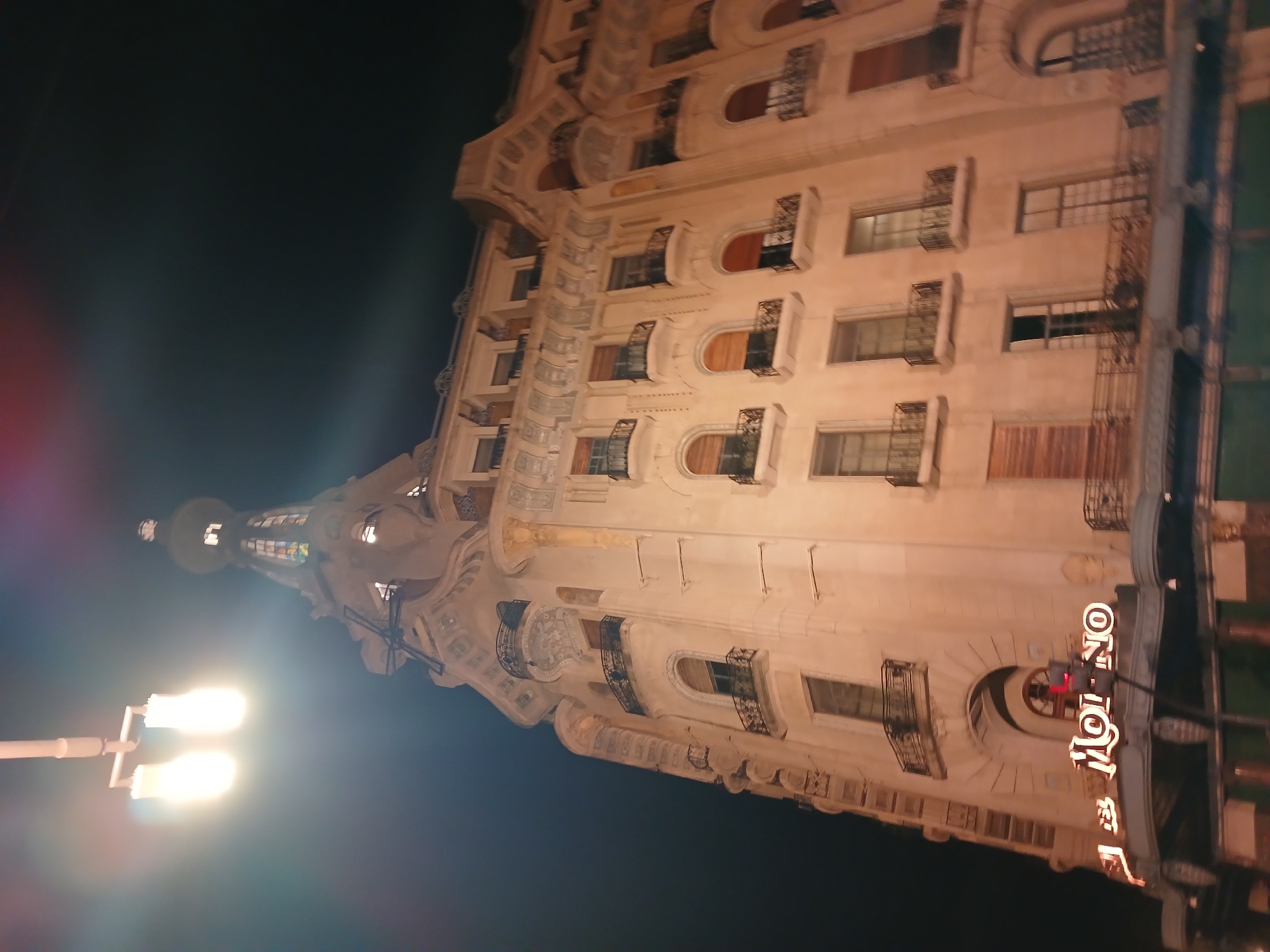 The image depicts the illuminated facade of the Confitería El Molino, located in Buenos Aires, Argentina, at night. The building is tall and ornate, featuring multiple floors with windows and balconies. The illuminated neon sign of "El Molino" is visible on the lower part of the building. A street light is seen on the left side of the image. The sky is dark.