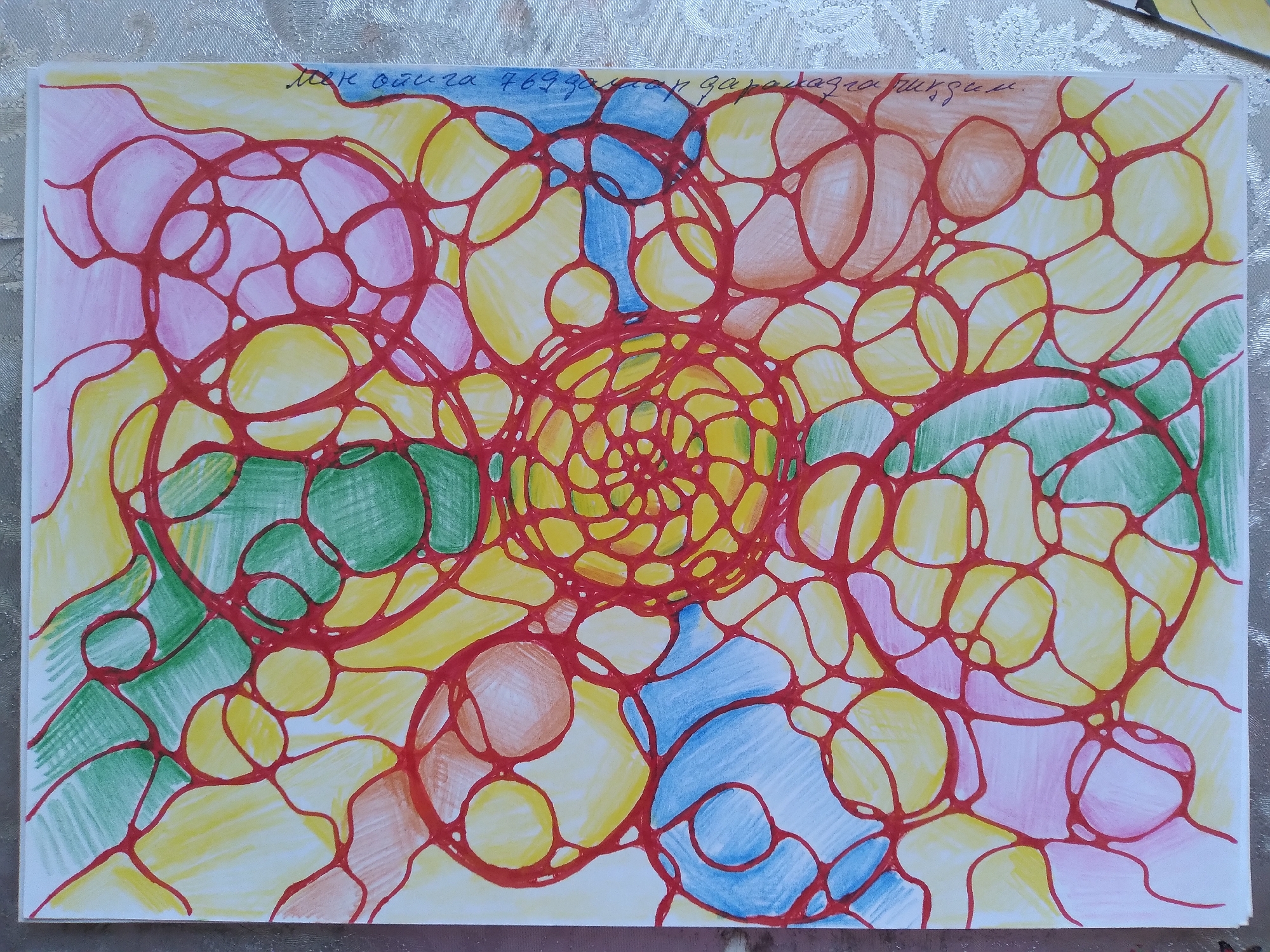 The image shows an abstract drawing on a white paper. The drawing is composed of interconnected red lines forming a network of circular and oval shapes. These shapes are filled with various colors, including yellow, pink, green, blue, and a light brown or orange. There is some text written in cursive script in the upper right corner of the paper, but it is illegible. The paper is lying on a textured white surface, possibly a tablecloth. The drawing does not depict any recognizable objects or scenes, and there is no indication of a specific location, including Baki, Uzbekistan.