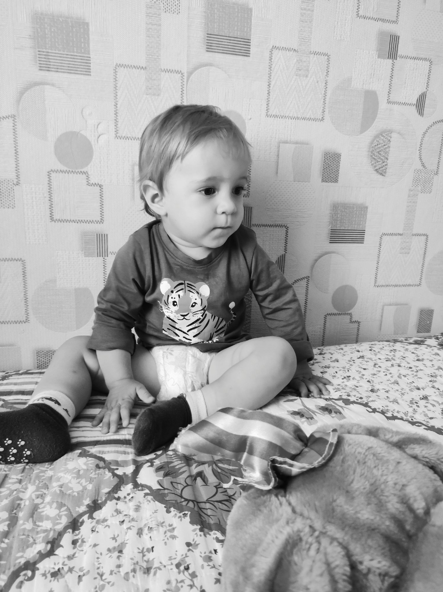 A young child sits on a bed or padded surface with a floral patterned blanket, in front of wallpaper featuring geometric shapes. The child is wearing a long-sleeved shirt with a tiger graphic and a diaper, and has socks on each foot. A stuffed animal lies on the bed to the right of the child. The image was taken in Chaikovsky, Russia.
