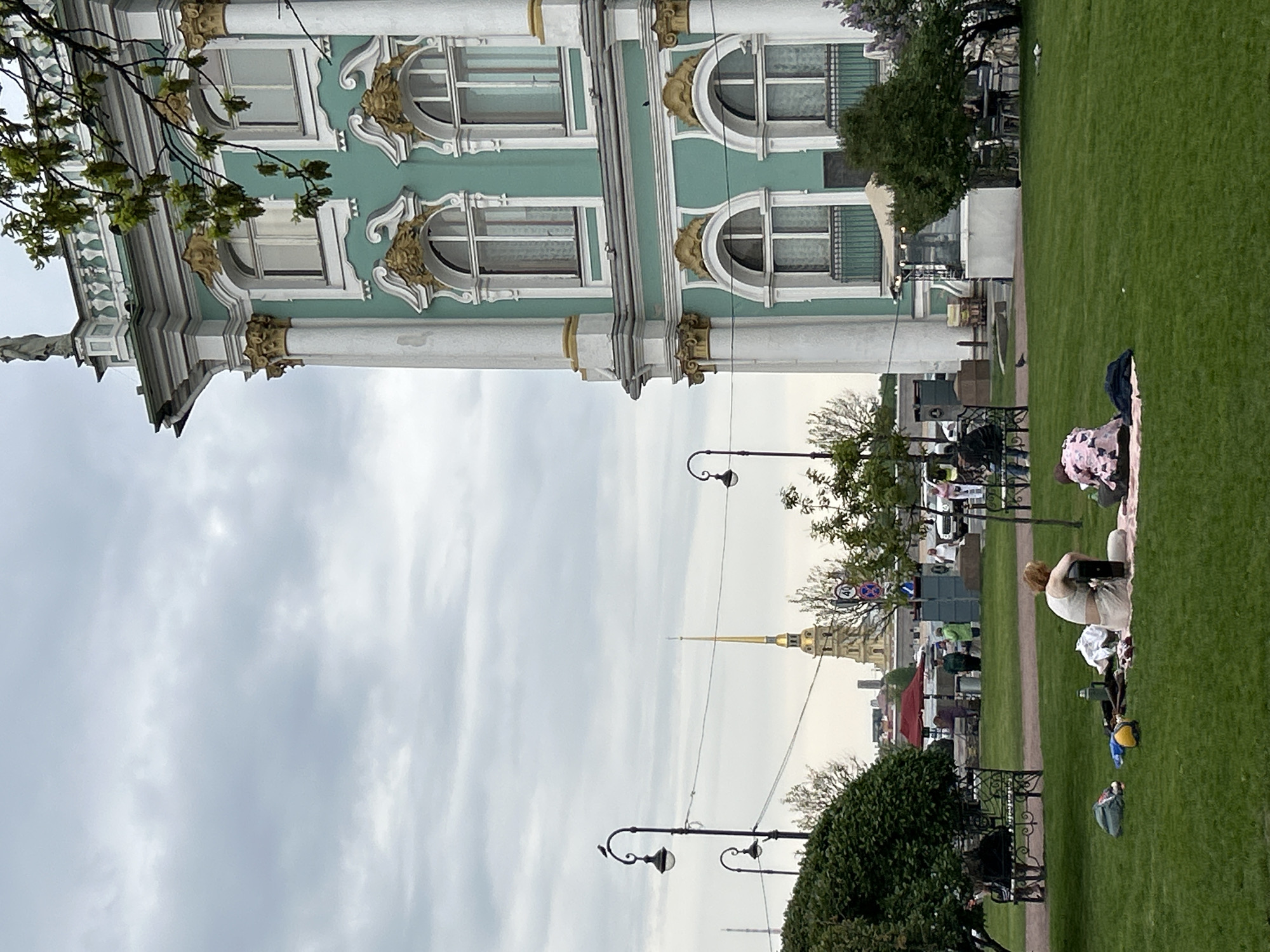 This daytime photograph captures a scenic view in Saint Petersburg, Russia, under an overcast sky. On the left, a grand architectural masterpiece, likely part of the Winter Palace or Hermitage Museum complex, dominates the frame. The building features a distinctive mint-green facade, adorned with white trim and elaborate golden architectural embellishments, including ornate window frames, columns, and decorative moldings. Tree branches with light green leaves partially obscure the upper left corner of the structure.
To the right, a large expanse of vibrant green lawn stretches across the foreground and midground, interspersed with a dirt path. In the immediate foreground on the grass, a person with reddish hair is seated, facing away from the camera, engrossed in a laptop or tablet. Various personal items, including bags and clothing, are spread around them on a blanket. Further back, several other individuals are visible throughout the park, some sitting on benches, others strolling along the path. There are also indications of outdoor cafes or market stalls in the midground.
In the distance, rising prominently above other buildings and trees, is the iconic golden spire of what appears to be the Peter and Paul Cathedral, a quintessential Saint Petersburg landmark. Ornate black street lamps with scrolled designs are visible in the park area, and power lines traverse the sky. The scene depicts a lively urban park setting adjacent to a historical monument. No clear text is discernible in the image.