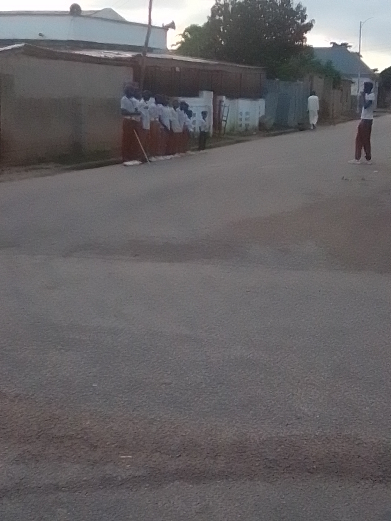 In Liji Hill, Nigeria, a group of individuals are standing in a line on a paved road. The individuals are dressed in white tops and red bottoms, suggesting they may be students or part of a formal group. To the right, another person is standing, wearing a white top and maroon pants, looking towards the other group. In the background, there are buildings with corrugated metal roofs and walls, typical of an urban or semi-urban environment in Nigeria. A lone figure in a white garment is walking away from the camera in the distance. The scene appears to be captured during the daytime, with an overcast sky.