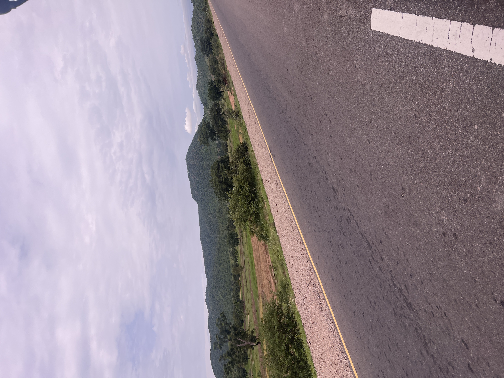 This image captures a rural roadside scene in Minda, Nigeria. The primary subjects are a paved road, a gravel shoulder with a yellow line, and a natural landscape of rolling, tree-covered hills and fields. The road occupies the right side of the frame, stretching into the distance. On the left, the landscape descends from the hills, revealing patches of green and brown fields interspersed with trees. The sky above is partly cloudy, suggesting it is daytime, likely late morning or early afternoon, with fair weather. There is no visible text in the image. The scene evokes a sense of journey and the natural beauty of the Nigerian countryside. The perspective, angled from the road, creates a feeling of movement or travel.
