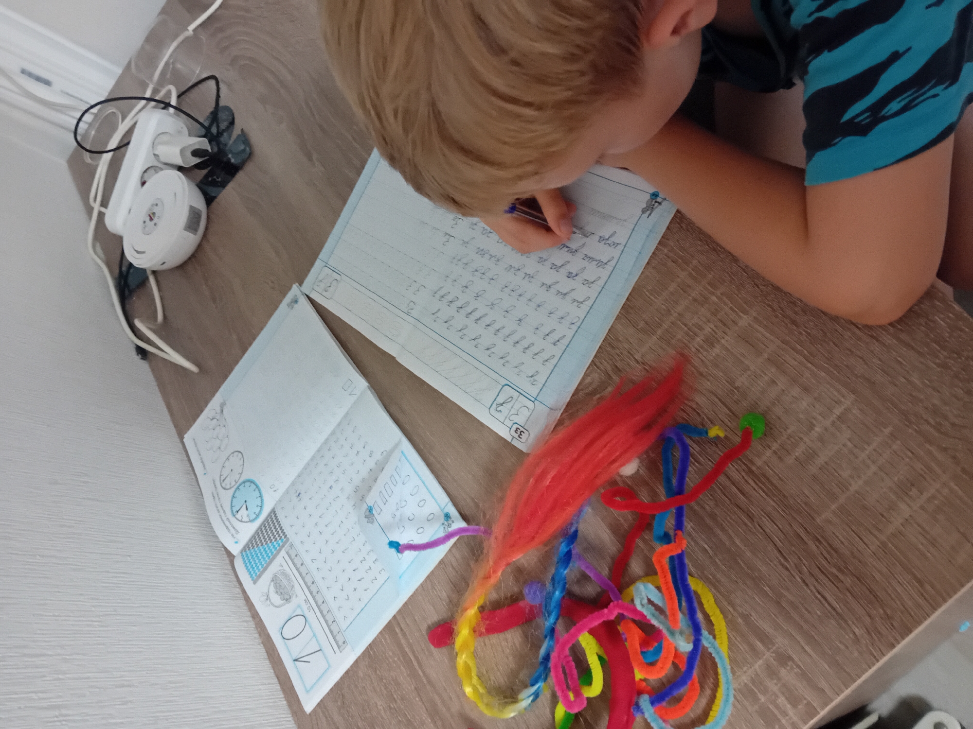 A young boy with blonde hair is sitting at a wooden desk, writing in a notebook with a blue pen. He is wearing a blue and black striped shirt. In front of him, there are two open notebooks. The left notebook has the number "10" on it, along with some math exercises and a ruler. The right notebook contains handwriting practice, with rows of letters and numbers. To the left of the notebooks, a colorful assortment of pipe cleaners and a red, fluffy material are scattered on the desk. In the background, on the right side of the desk, a white power strip with several plugs and cords is visible, along with a white round object, possibly a router or speaker. The wall behind the desk is white. This image appears to be taken indoors in a residential setting.