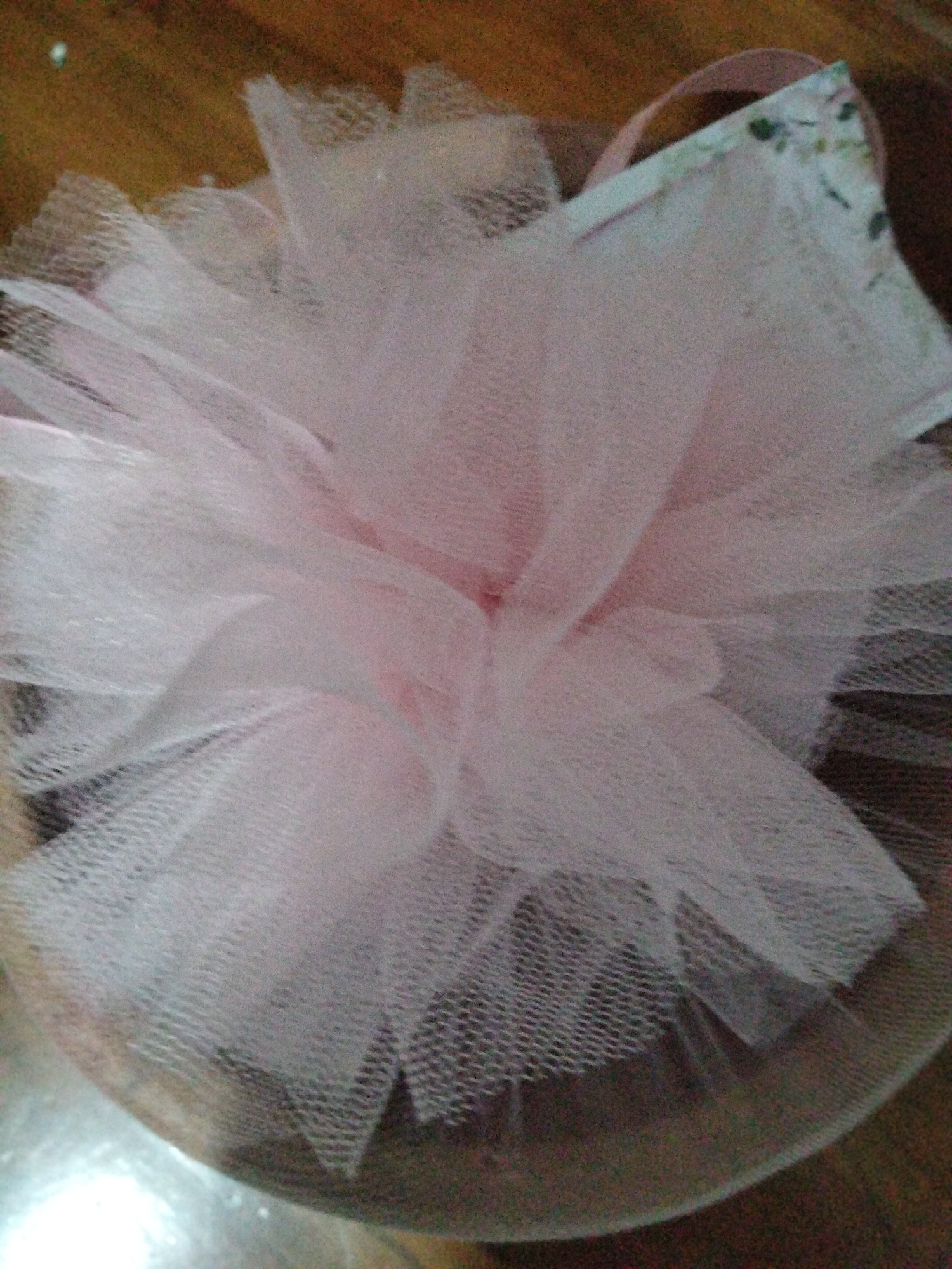 The image shows a close-up, overhead view of a pink tulle fabric gathered and arranged in a fluffy, layered fashion, resembling a flower or a pouf. The fabric is translucent, with a visible mesh pattern. It is situated on a wooden surface, and part of a box with a floral pattern and a pink ribbon is visible behind the tulle. The lighting is dim, and the image quality is poor, with a significant amount of noise and blur. The scene does not depict any identifiable landmarks or elements that would allow for the determination of its location within Payagaboh, Indonesia.