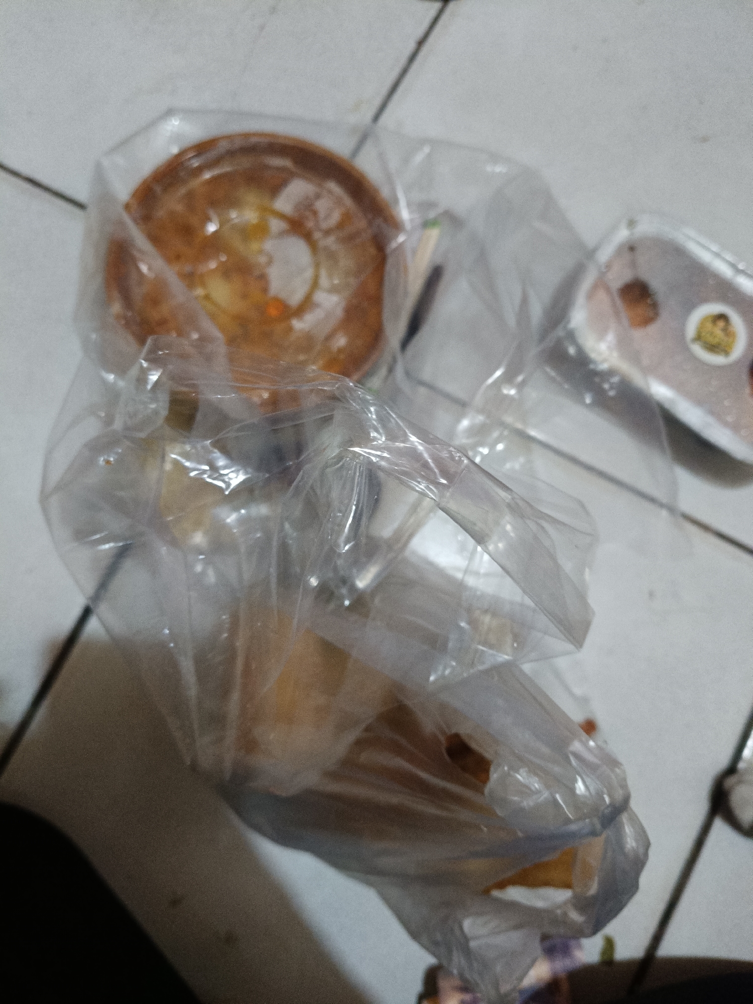 In Cileungsi, Indonesia, a transparent plastic bag containing multiple food items rests on a light-colored tiled floor with dark grout lines. Prominently visible within the bag is a round, golden-brown baked item, possibly a cake or pie, showing a textured top. Other lighter-colored food items, consistent with pastries or bread, are also contained within the bag. To the right, a separate clear plastic container with a white base holds dark-colored contents and features a circular sticker on its lid. No human figures or active interactions are visible in the scene.