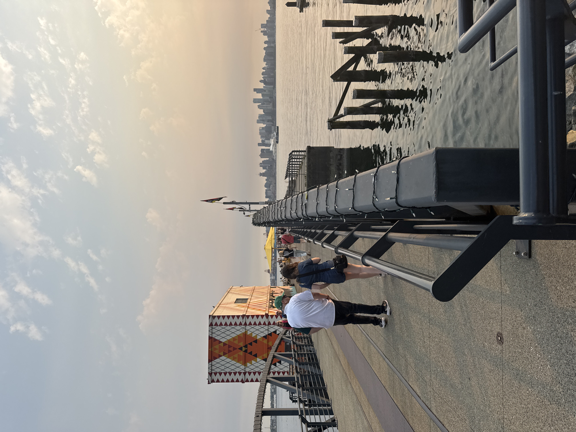 This image shows a view of the waterfront in North Vancouver, Canada, at what appears to be sunset. The foreground features a concrete pier with a railing adorned with string lights. Several people are walking on the pier. In the mid-ground, a distinctive orange and white patterned building is visible. The water of the Burrard Inlet is to the right, with several weathered wooden pilings protruding from it. In the distance, the Vancouver skyline is clearly visible across the water. The sky is a soft, warm hue, indicating late afternoon or early evening light.