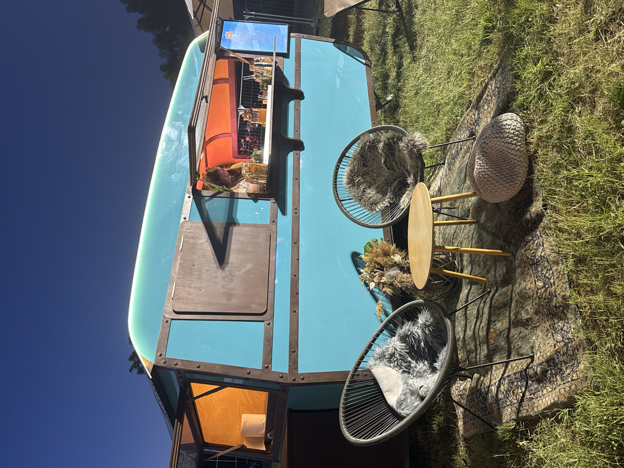 A turquoise and brown food trailer is set up outdoors on grass at night. The trailer has an open window with a person behind the counter, serving drinks. In front of the trailer are two dark, wire-frame chairs with fluffy grey cushions, a small wooden coffee table, and a light-colored knitted pouf, all arranged on an ornate rug. Dried floral arrangements are placed near the chairs. The background shows a dark sky and trees. The image was taken at the "Soul Kitchen" event in Sofia, Bulgaria.