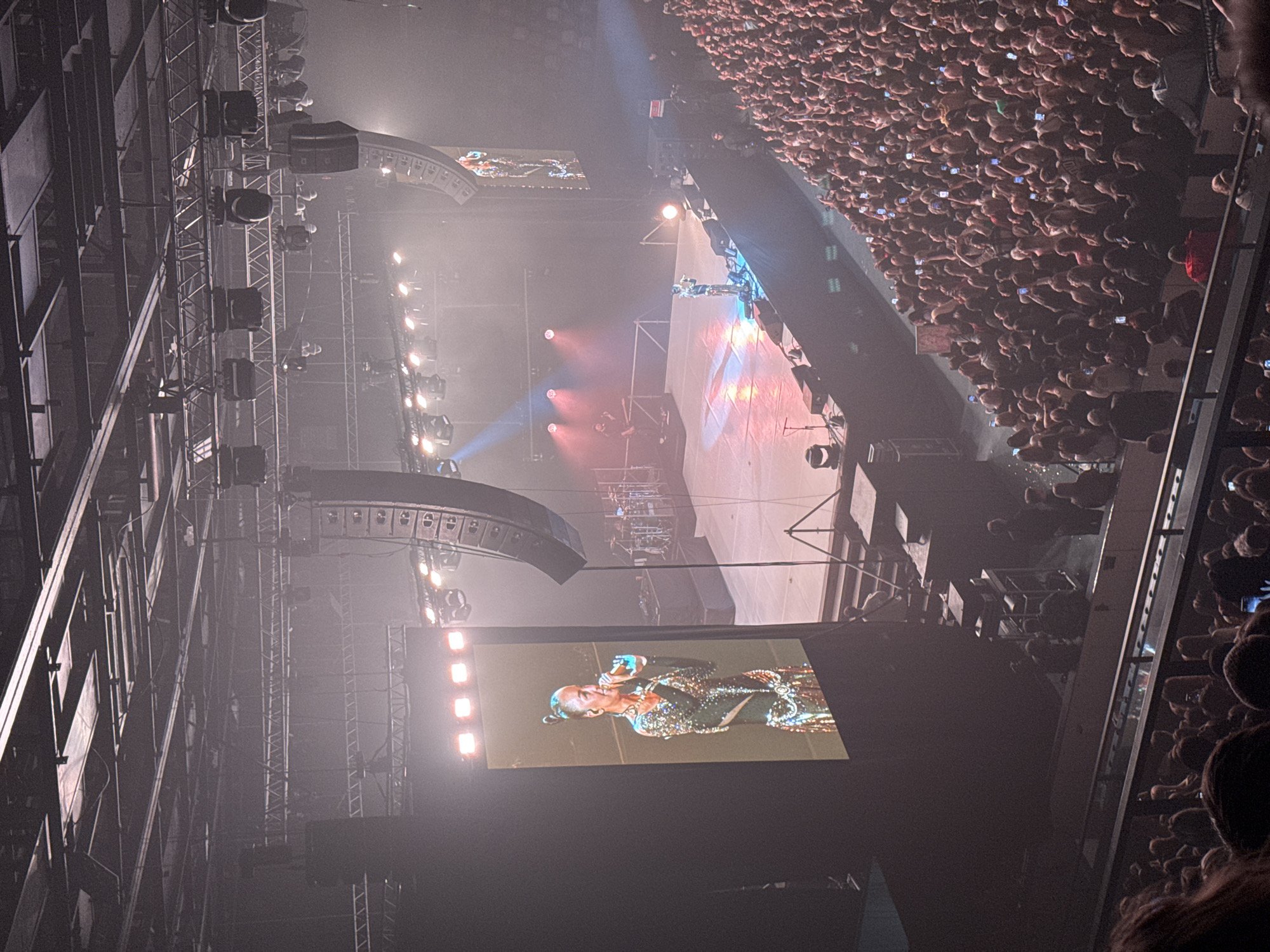 This vertical image captures an expansive view of a large-scale indoor concert or performance in Perm, Russia, taken from an elevated perspective, likely from an upper-level seating area. The scene is dominated by a brightly lit stage area on the left (when viewed horizontally) and a massive, densely packed audience filling the right side of the frame.
On the main stage, a lone performer, small in comparison to the venue, is visible. Above and around the stage, a complex network of metal trusses, speaker arrays, and numerous spotlights creates a dramatic lighting display, with beams of light piercing through a hazy atmosphere. Red and blue stage lights illuminate parts of the stage and surrounding structures.
Two large video screens are prominent. One, closer to the foreground, displays a close-up of a female performer. She has her hair styled in a bun, is wearing an intricately sparkling, ornate costume, and is holding a microphone. Another, smaller screen further back on the stage, shows a wider view of the performance or additional visual content.
The audience, a sea of heads, extends far into the background. Many individuals in the crowd are holding up their illuminated mobile phones, likely recording the event or taking photos, indicating a high level of engagement and excitement. The setting is clearly an evening or night event, given the reliance on artificial lighting. The overall impression is one of a major, energetic live performance with a large attendance. No explicit text is visible in the image itself, beyond the imagery on the video screens.