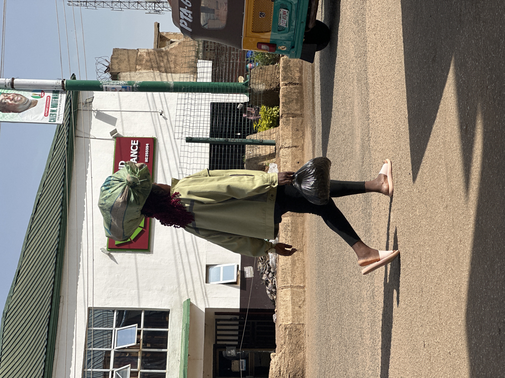 A woman walks across a paved road in Bukuru, Nigeria, carrying a large green sack balanced on her head and a black plastic bag in her right hand. She wears a light green jacket, dark leggings, and pink sandals. The road is lined with a concrete curb and a sidewalk with some vegetation and debris. Behind the sidewalk, a white building with a green roof and large windows is visible, along with a utility pole with a political banner. A yellow and green auto-rickshaw is parked on the right side of the road. The sky is clear and blue.