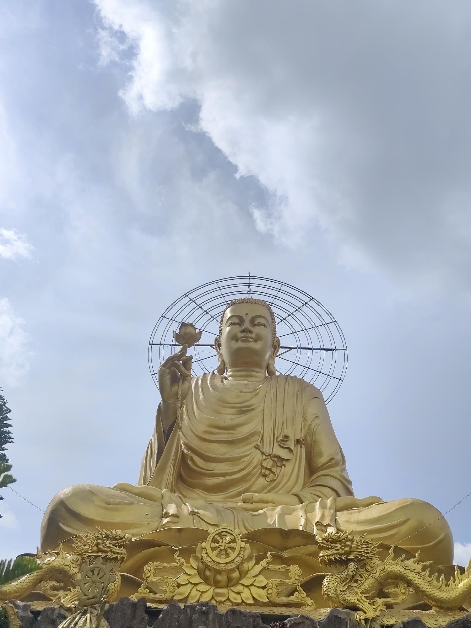 A low-angle, medium shot captures a golden Buddha statue situated in Lâm Viên - Đà Lạt, Vietnam. The statue, posed in a seated, meditative position, holds a lotus flower in its left hand. A circular, wire-frame structure forms a halo-like design behind its head. The base of the statue is adorned with golden dragons and decorative elements. The sky, partially cloudy, serves as the backdrop. Green foliage is subtly visible on the left edges of the frame.
