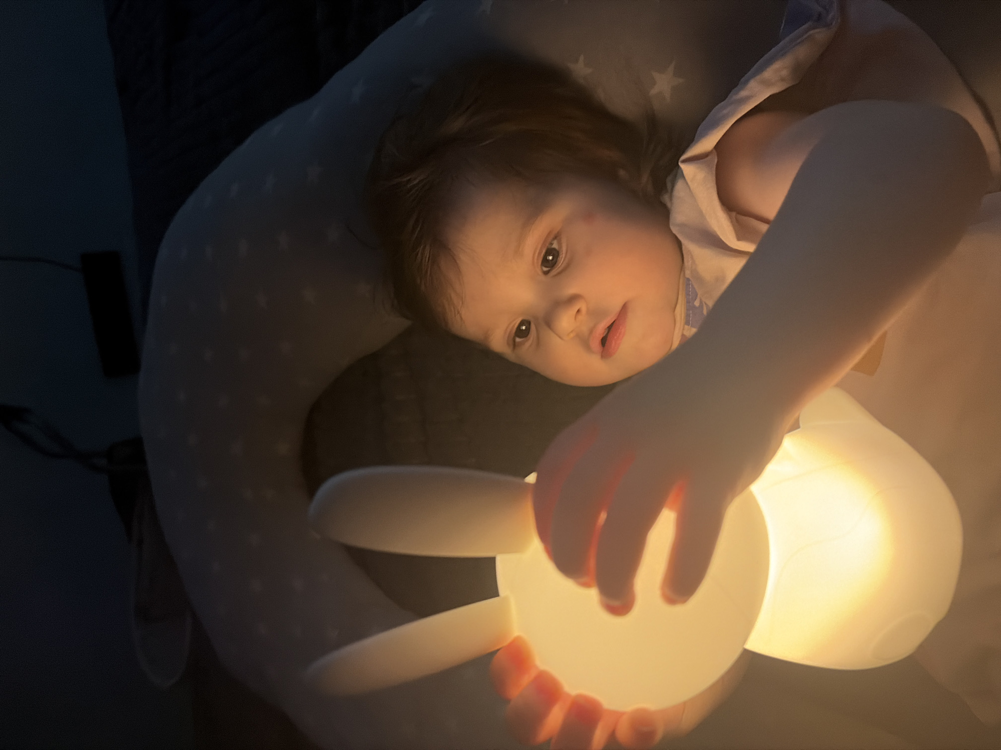 A baby girl, lying on her back, is illuminated by a glowing bunny-shaped lamp held in her hands. She is on a star-patterned pillow. The lighting is dim, indicating an indoor setting, likely a bedroom. The image does not provide any information about the location in the city of Chaikovsky, Russia.