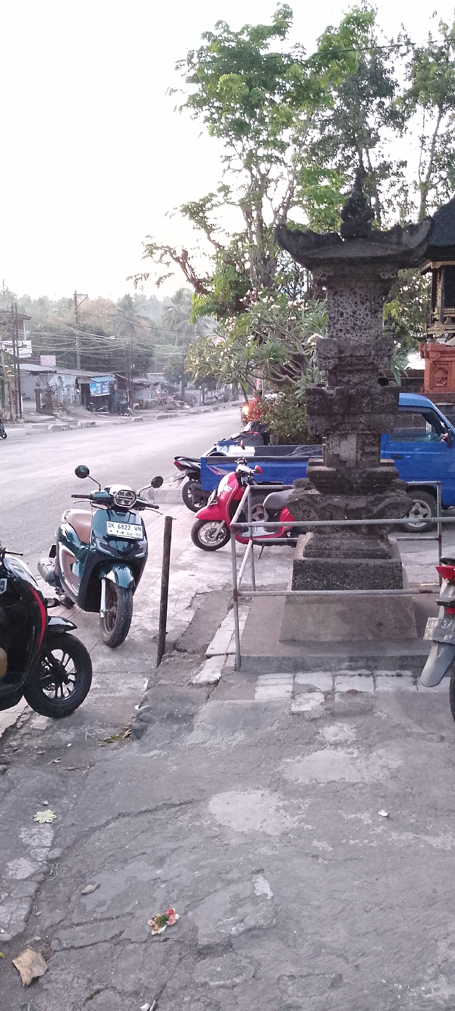 The image depicts a street scene in Pekutatan, Indonesia. In the foreground, there are several parked scooters. A dark-colored scooter is partially visible on the left. To its right, a teal-colored scooter with a brown seat is prominently displayed, bearing the license plate "DK 6822 HN". Behind the teal scooter, a red scooter is parked. Further back, a blue pickup truck is visible. To the right of the scooters and truck is a stone structure, possibly part of a temple or monument, adorned with decorative elements. In the background, there is a street lined with buildings and utility poles with numerous wires. Trees with green foliage are visible throughout the scene, with some bearing white flowers. The ground is paved with asphalt and concrete, showing signs of wear and tear. Scattered on the ground are a few leaves and what appears to be a small floral offering. The overall lighting suggests it is daytime, possibly late afternoon or early morning due to the soft light.
