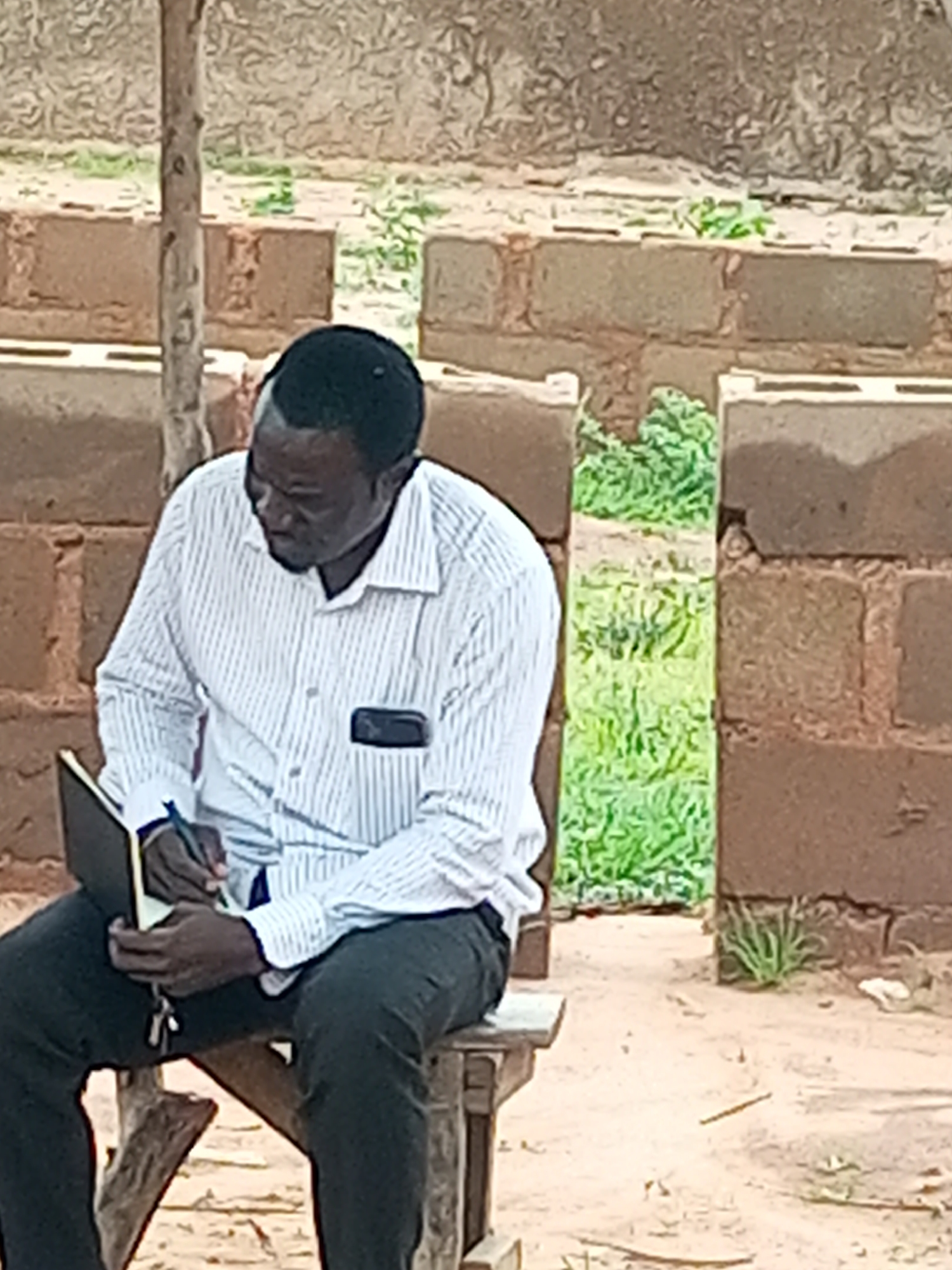A Black man, wearing a white pinstripe shirt and dark pants, is seated on a wooden stool, writing in a notebook. He is in a dimly lit outdoor environment with a brick wall and vegetation in the background. The scene appears to be in Tempure, Nigeria.