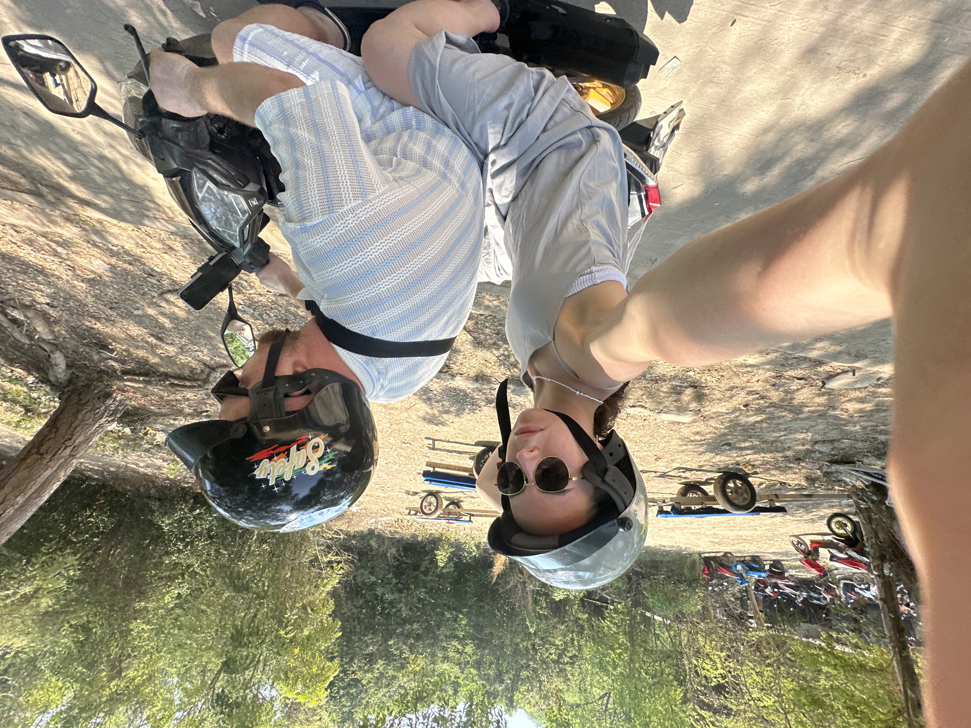 A couple is pictured from a low angle, sitting on a black scooter in an outdoor parking area. The man on the left wears a black helmet and a short-sleeved, light blue and white striped shirt. The woman on the right wears a gray helmet and sunglasses, and a gray short-sleeved shirt. The couple is presumably taking a selfie. Behind them are trees, trailers, and many parked scooters. The location is Pattaya City, Thailand.