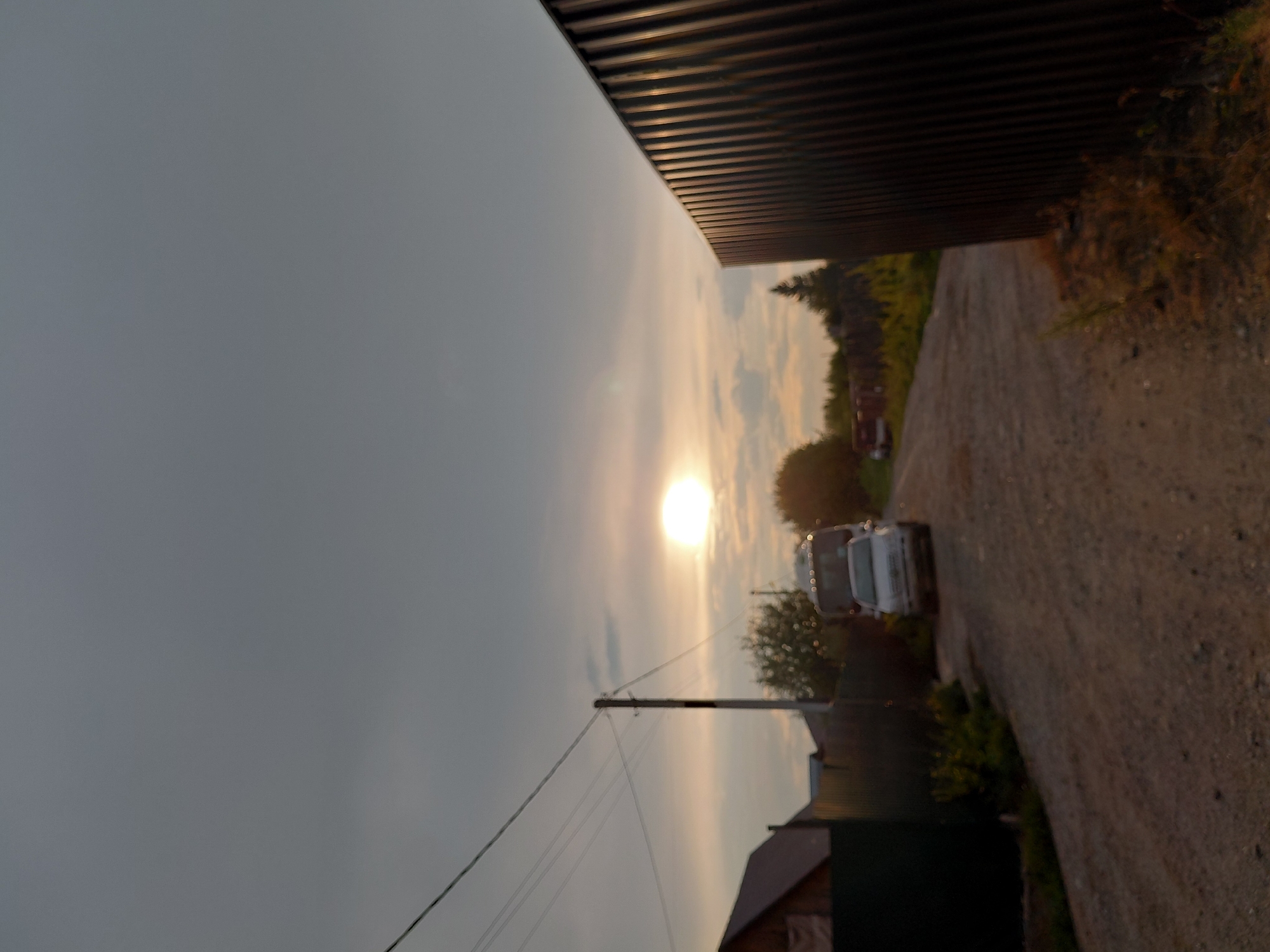 The image depicts a rural or suburban street scene in the city of Novosibirsk, Russia, during sunset. The sky is overcast with a bright sun low on the horizon, casting a warm glow. A gravel road occupies the foreground and midground, with a dark, corrugated metal fence on the right side of the frame. To the left, a power pole with multiple wires is visible, along with the roof of a building. Further down the road, a light-colored passenger car is parked, and behind it, a white van or small bus is also present. Green vegetation and trees line the sides of the road, and another fence is visible in the distance. The overall impression is one of a quiet, late afternoon or early evening in a residential area.