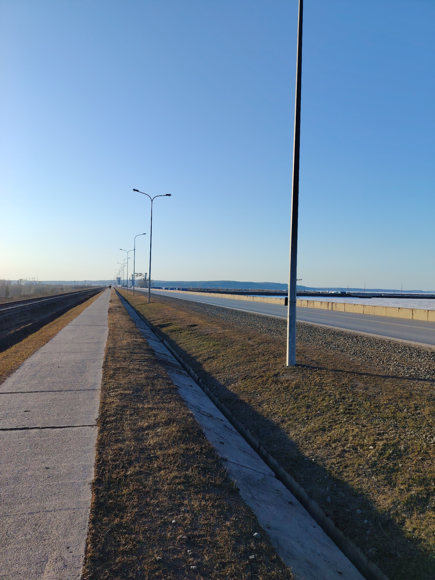 The image depicts a section of a pathway and road in Chaikovsky, Russia, under a clear blue sky. A gray concrete pathway runs along the left side of the image, bordered by dry grass and a drainage ditch. Adjacent to the pathway is a road with a gray gravel shoulder. The road is partially obscured by a concrete barrier. A row of streetlights lines the pathway and road. A tall utility pole stands on the right side of the frame. In the distance, there is a body of water and some industrial structures. The lighting suggests it's either early morning or late afternoon.