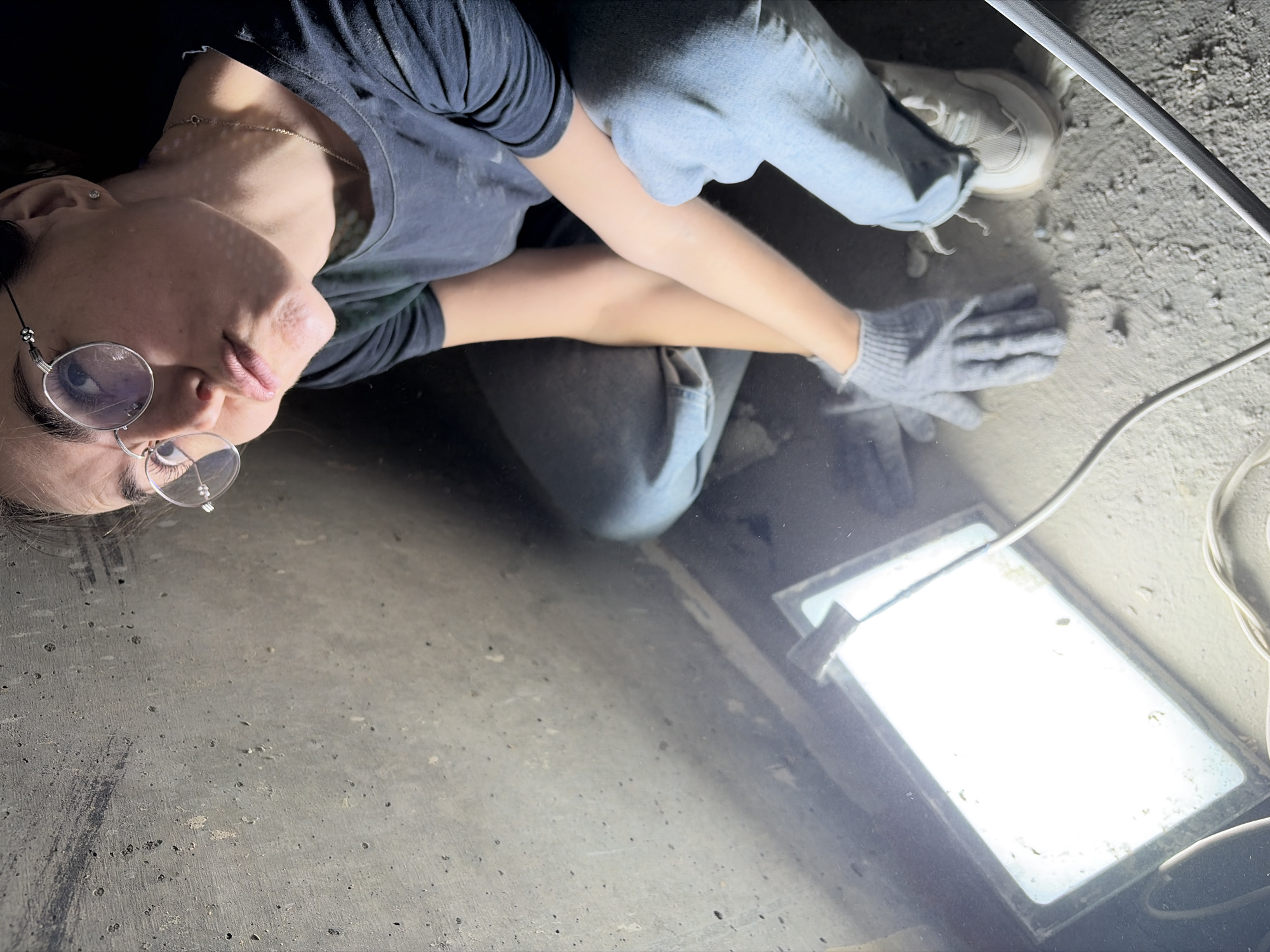 This overhead image shows a young woman situated in an indoor utilitarian setting. She is positioned on her side, looking upwards towards the camera with a neutral expression, her head tilted slightly. She wears a dark t-shirt, light blue jeans, and white sneakers. A silver chain necklace and a small ear stud are visible. Round, wire-framed glasses are perched on her nose.
Her right arm, covered by a grey knitted glove, rests on a dark, highly reflective surface, possibly a polished floor or a large pane of glass, which mirrors her form and the bright light source. To her right, a rectangular, intensely bright light fixture, likely a work lamp or LED panel, illuminates the scene and is also reflected by the surface. White electrical cables are visible trailing around the light and across the surrounding floor.
The floor around the reflective area is made of concrete, appearing textured, dusty, and speckled with small debris. The posture of the woman and the presence of the work light, gloves, and concrete suggest she might be engaged in a task or inspection on the ground, possibly in a workshop, construction area, or basement. The artificial light makes the time of day indeterminate. There is no visible text within the image. While the location context is Izhevsk, Russia, no specific visual cues in the image confirm this.