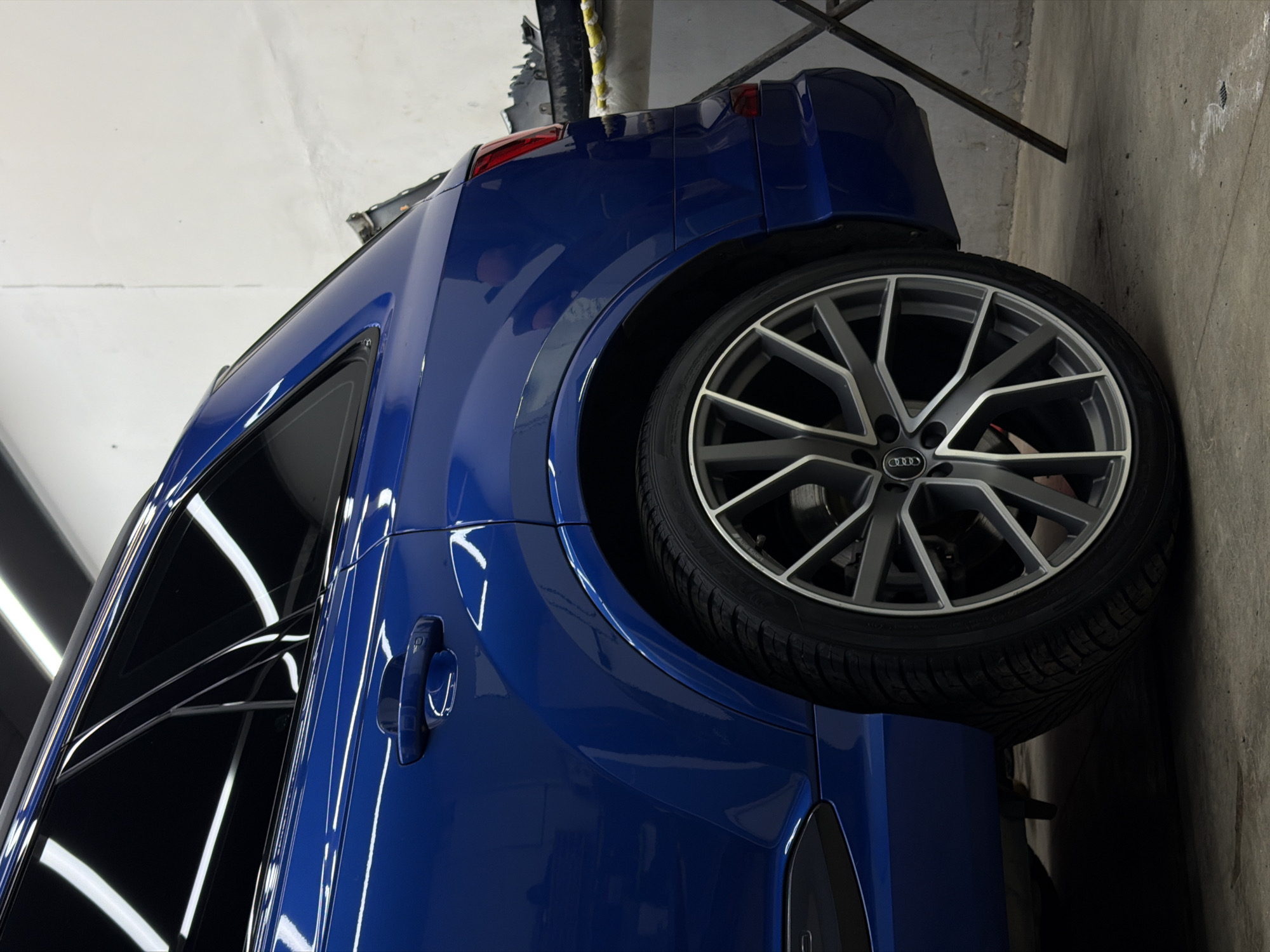 A glossy blue Audi car, likely an SUV or crossover, is prominently featured, parked indoors. The image is captured with a 90-degree clockwise rotation, presenting the vehicle as if resting on its side against a concrete surface.
The car's vibrant, deep blue paintwork is highly reflective, showcasing linear artificial light sources, particularly on the doors, rear quarter panel, and windows. The rear passenger door handle is visible, matching the car's body color. The side windows are tinted, further enhancing the reflections of the overhead lighting.
A large, multi-spoke alloy wheel, featuring a stylish two-tone design with dark grey spokes and polished accents, is a significant detail. The "Audi" logo is distinctly visible in the center cap of the wheel. The tire displays a robust tread pattern.
The background consists of plain concrete walls or floor, and some dark metallic structures or racks are partially visible, suggesting an industrial or workshop environment, such as a detailing studio or garage. No individuals are present in the image. The clean, pristine appearance of the car implies it may have recently undergone cleaning or detailing.
