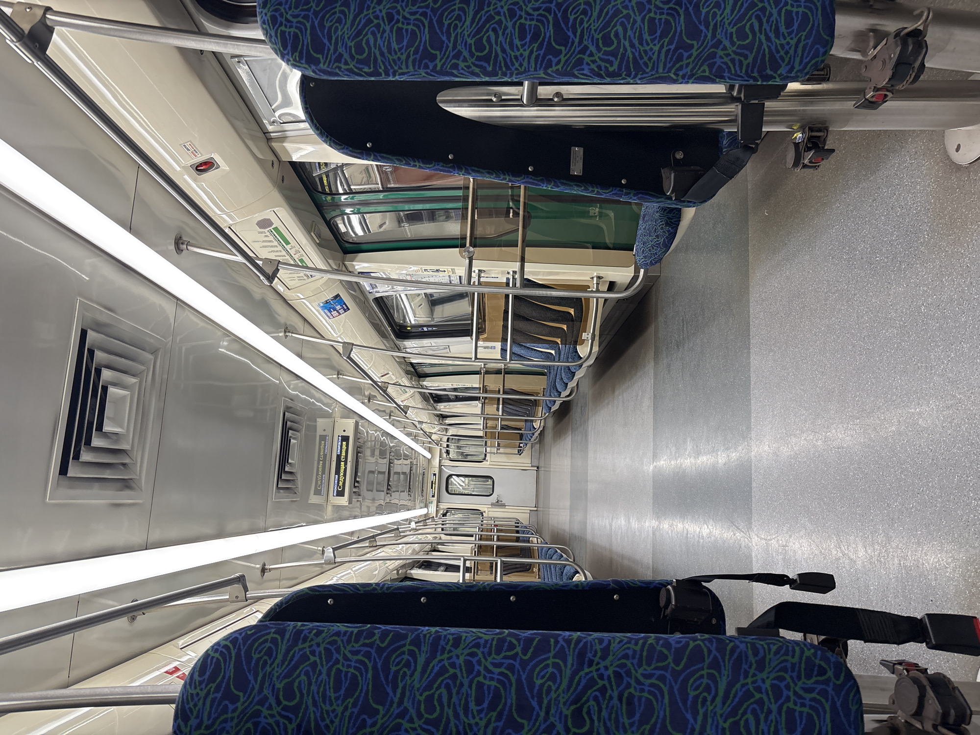 The image shows the interior of a modern subway car with blue and green patterned seats. The car appears to be empty. Overhead lights illuminate the carriage, and a ventilation grate is visible on the ceiling. A sign with Russian text, indicating "Next station," is displayed above the seats. The floor is a speckled grey material. The overall environment suggests a public transportation setting, likely a subway or train, designed for passenger travel. The image was taken in Novosergievka, Russia.