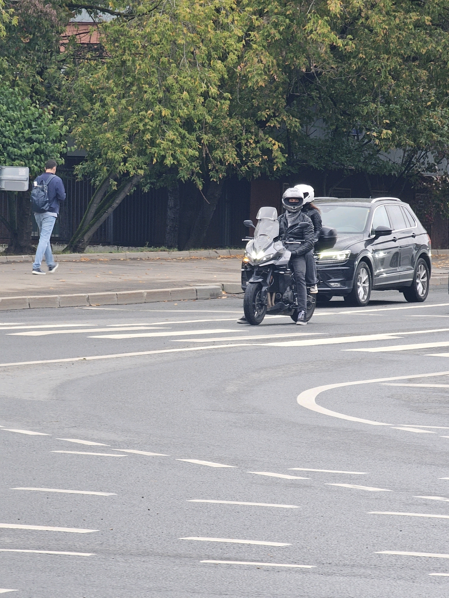 An overcast day in a city, likely Moscow, Russia, features a wide asphalt road with white lane markings. On the right side of the road, a black SUV is parked or stopped. To the left of the SUV and also on the road, a motorcycle with two occupants is positioned. The driver of the motorcycle is wearing a black leather jacket, dark pants, and a white helmet with a dark visor. The passenger, also wearing a white helmet and dark clothing, is seated behind the driver. In the background, a pedestrian wearing a blue jacket and backpack is walking on a sidewalk bordered by trees and a dark fence. The environment suggests a street scene with traffic and pedestrian activity.