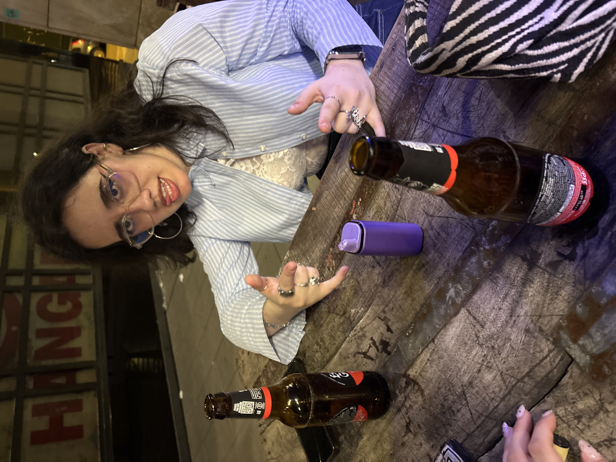 The scene depicts a woman seated at a wooden table in what appears to be a bar or restaurant. The location is likely in the city of Córdoba, Spain, as there's a reflection of what appears to be a sign for "Hanga" in the background, potentially a restaurant there. The woman has dark hair, wears glasses, a blue striped shirt, and several rings on her fingers. On the table, there are two brown beer bottles, a purple vape pen, and an adjacent table with a zebra-print cushion. Another person is partially visible with their hand at the lower right side of the image, showing painted fingernails. The overall environment is dimly lit, suggesting it's either evening or the interior of a dimly lit establishment.