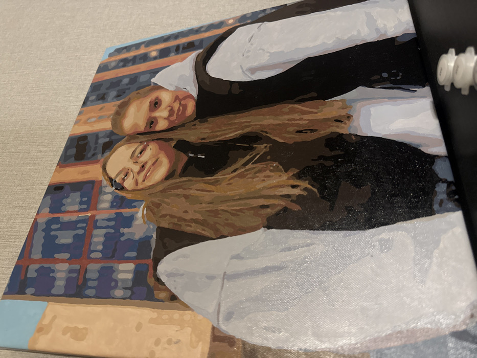 The image displays a photograph of a stylized canvas painting, oriented diagonally, resting on a dark surface and leaning against a textured, light-colored wall. In the lower right foreground of the photograph, three small, white cylindrical containers with caps are stacked vertically.
The painting itself depicts two young adult individuals, likely women, in a close-up, selfie-style composition. The individual on the left has long, light brown hair, wears glasses, and a dark long-sleeved top with a visible zipper detail, smiling towards the viewer. Their left arm, covered in a white sleeve, extends forward, contributing to the wide-angle perspective. The individual on the right has shorter, light brown hair and wears a dark sleeveless vest over a white long-sleeved hooded sweatshirt, also smiling gently towards the viewer.
The background of the painting features stylized urban architecture: a multi-story building rendered in orange-brown tones with multiple rectangular windows, some displaying blue hues and others darker tones. To the left of this building, a section of a lighter orange-beige wall is visible under a segment of light blue sky. These elements depict a generic city environment. While the location context of Krasnodar, Russia is provided, no specific identifiable landmarks or unique architectural features are present in the stylized artwork to pinpoint an exact location within the city.