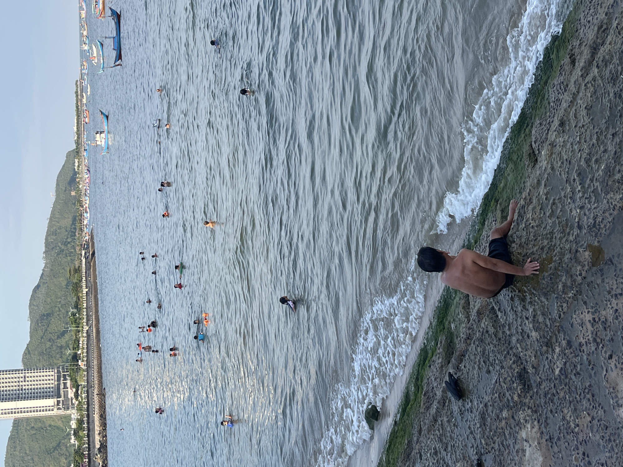 This image captures a vibrant coastal scene in Bac Nha Trang Ward, Vietnam. In the foreground, a shirtless man wearing dark shorts sits on a textured, dark gray rocky shoreline, which is partially covered in green algae and wet from the ocean. He is positioned with his back to the camera, looking out at the water. A dark object, possibly footwear, rests on the rock beside him.
The ocean water, exhibiting moderate ripples and small breaking waves near the shore, contains approximately 20-30 individuals engaged in swimming, wading, and floating. Several people are visible wearing orange or dark life vests, and some are using orange inflatable rings or floats. One person closer to the shore is swimming on their back, while another wears a blue swimming cap and goggles.
In the background, a prominent tall, modern white building with numerous windows stands against a backdrop of lush green hills and mountains. A paved promenade or road with streetlights and a low wall separates the land from the water. Further out in the bay, a considerable number of small, colorful fishing boats, primarily blue with red and yellow accents, are moored or drifting. The sky is a light blue with some atmospheric haze.