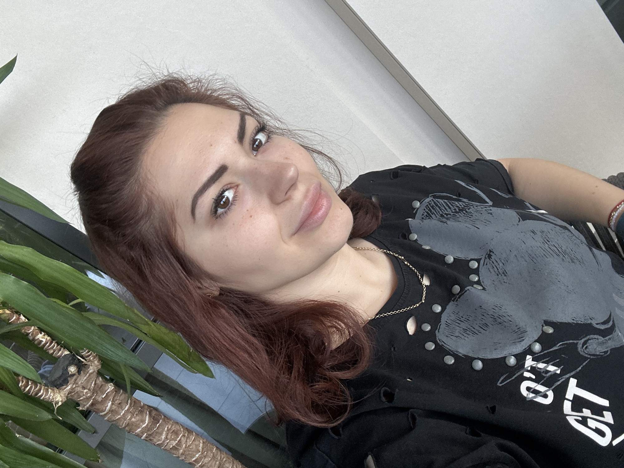 A woman is seen from a low angle. She has light skin, brown hair, and brown eyes. She is wearing a black t-shirt with a grey graphic on it, as well as a silver necklace. The background consists of a white wall and what appears to be a green plant. The photograph may have been taken indoors. The location is not evident from the image.