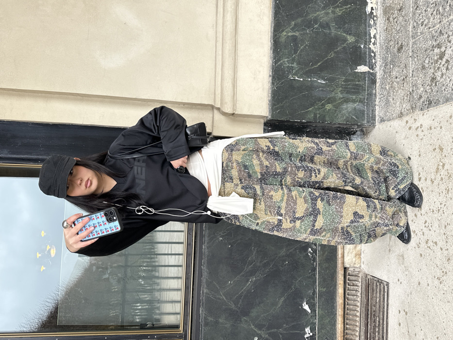 A young female, appearing to be of East Asian descent, stands facing the camera on a light grey concrete sidewalk in Paris, France. She wears a black long-sleeved V-neck top with dark stylized text, possibly "ONERA," across the chest. A white garment is wrapped around her waist, tied in the front. Her lower body is clad in baggy, wide-leg camouflage pants with a textured, mottled pattern of green, brown, and beige. She wears black flat shoes. A black shoulder bag hangs over her left shoulder. She has long, dark hair and wears a black cap and a silver ring on her right index finger.
In her right hand, extended towards the viewer, she holds a light blue iPhone with a patterned case displaying small, repeating red stylized icons. White wired in-ear headphones hang from the phone, with the wires loosely bundled around her neck. Her left hand is partially tucked into her waistband. She maintains a neutral expression, looking directly forward.
The subject is positioned against a building facade. To her left, a large rectangular window with a dark frame reflects bare tree branches, suggesting a Parisian street scene and an overcast sky. Further reflections in the window suggest internal lighting fixtures or elements from across the street. Below the window and spanning the lower portion of the building is dark green marble paneling, exhibiting some visible wear and white scuff marks. To her right, the facade transitions to a lighter, possibly cream-colored stone or stucco section with a horizontal decorative molding. A metal grate is visible on the sidewalk to her left. The architectural style is consistent with typical urban buildings found in central Paris.