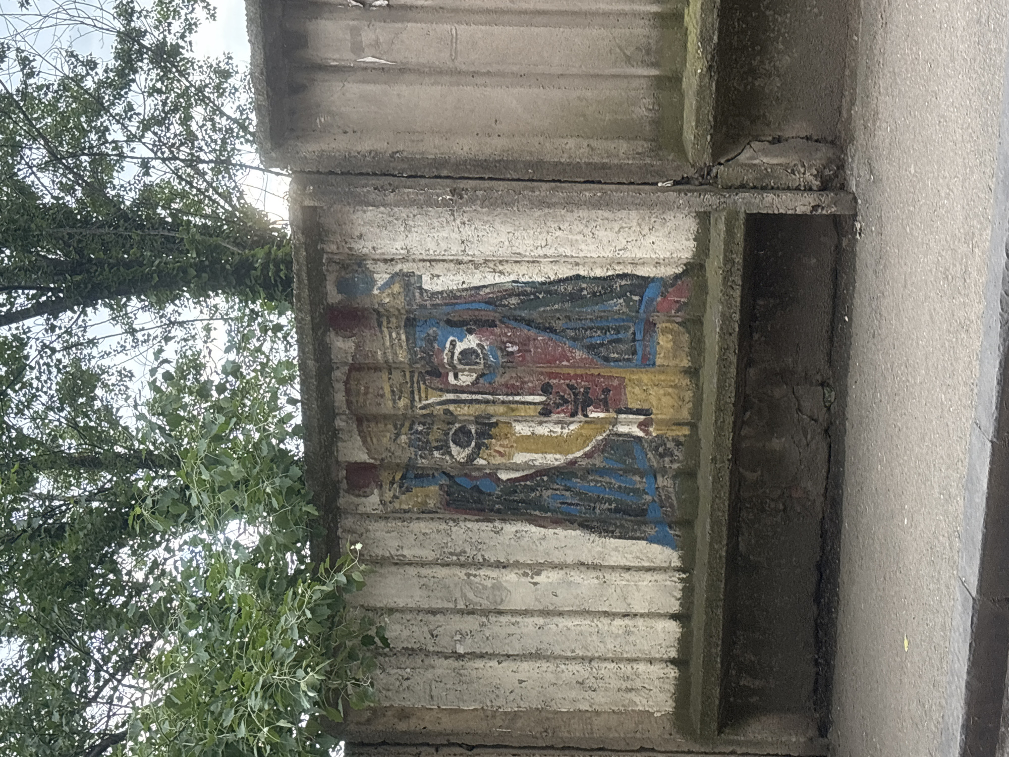 A segment of an aged, concrete structure supports what appears to be an eroded painting of two figures. The figures are depicted in a stylized, possibly religious or historical, manner, with discernable details suggesting crowns or headdresses and flowing garments. The painting's colors are faded, with reds, blues, and yellows showing through the wear. Flanking the concrete structure on the left is a tree with green foliage. To the right of the concrete structure is a concrete wall. The scene is located outdoors. Based on the style of the artwork, and the concrete structure, it is possible this image was taken in Tbilisi, Georgia.