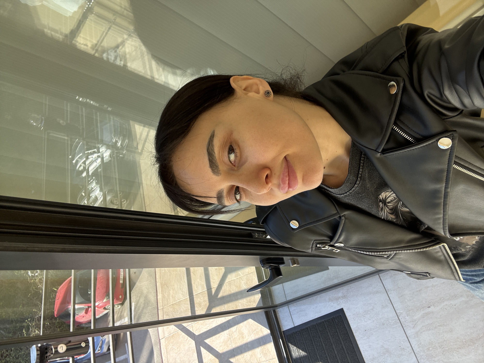 A woman with dark hair and fair skin stands facing the camera, wearing a black leather jacket over a gray patterned t-shirt. She is positioned indoors, near a window or glass barrier that reflects an outdoor scene.
Outside, a red and blue vehicle can be seen through a metal railing, parked on a tiled surface. The location is likely an urban setting.
Without additional clues or metadata, it is not possible to confirm the specific location in Tbilisi, Georgia.
