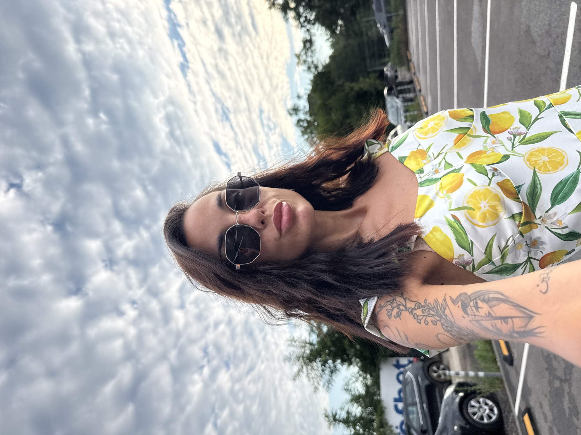 A woman with dark, wavy hair and sunglasses takes a selfie outdoors in a parking lot. She is wearing a white dress with a lemon print. Her left arm, visible on the left side of the frame, is adorned with a large tattoo depicting a face with horns and floral elements. In the background, trees and cars are visible. The sky above is cloudy. The scene appears to be located in the city of Pleven, Bulgaria.