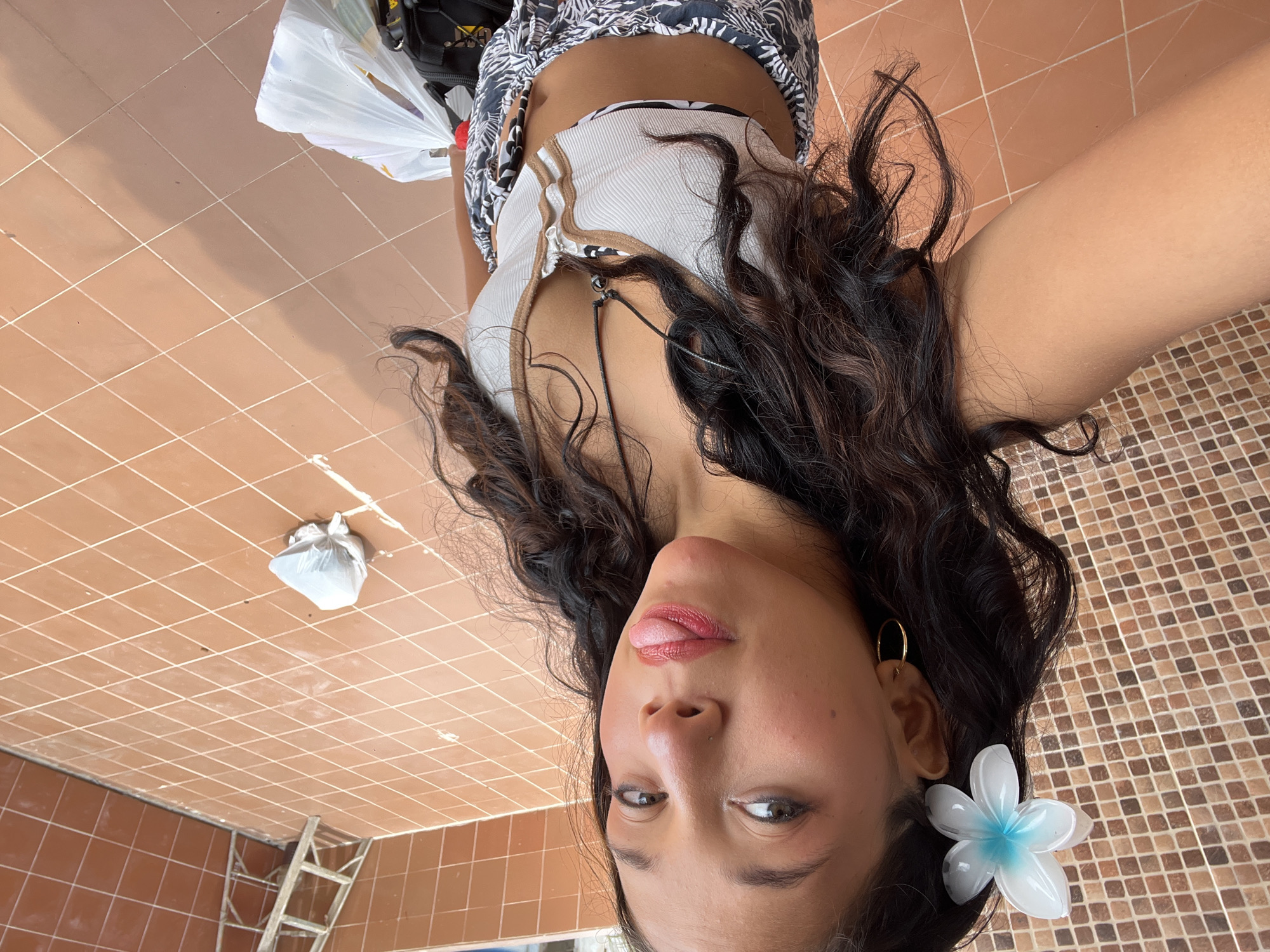 A woman is taking a selfie, her face close to the camera with her tongue slightly out. She has long, dark wavy hair, an earring, and a white flower in her hair. She is wearing a white top with brown trim and a black and white patterned skirt. A black bag and a white plastic bag are visible behind her. The background consists of brown square tiles on the floor and wall. A ladder is visible in the lower-left corner. Pampatar, Venezuela, is not evident in the image.