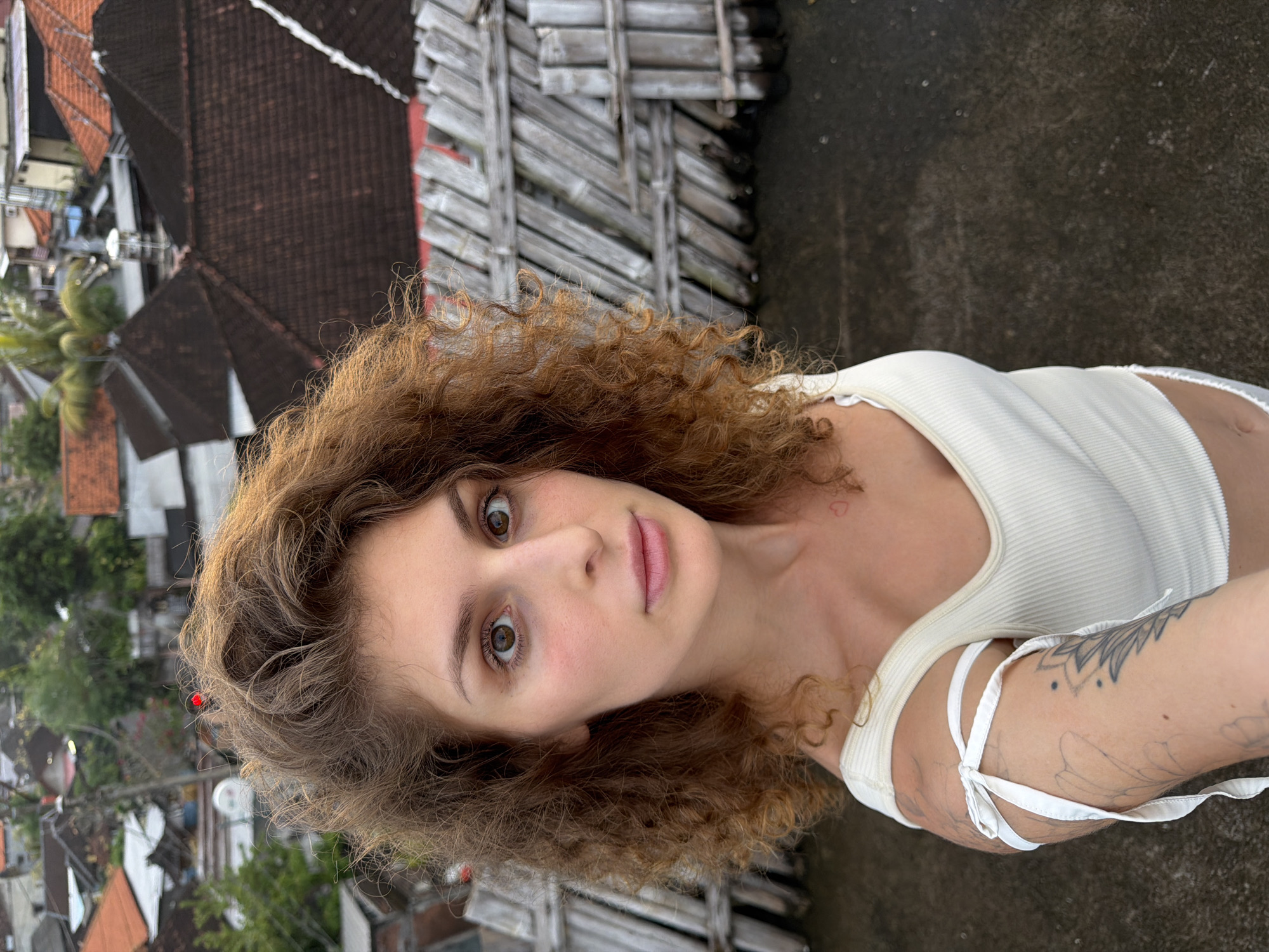 The image shows a young Caucasian woman with curly auburn hair taking a selfie. She is wearing a white ribbed tank top with white ribbon ties on the shoulders. She has tattoos on her left arm, including a mandala design. A small red heart tattoo is visible on her collarbone. Her makeup is minimal, and she has brown eyes. The background suggests an outdoor urban environment, likely a rooftop or elevated area, with a view of many tiled roofs, palm trees, and various buildings, consistent with the cityscape of North Kuta, Indonesia. A weathered wooden fence structure is prominent in the mid-ground to the right of the subject. The lighting indicates it is daytime, possibly late afternoon or early morning given the soft light. The woman is looking directly at the camera with a neutral expression.