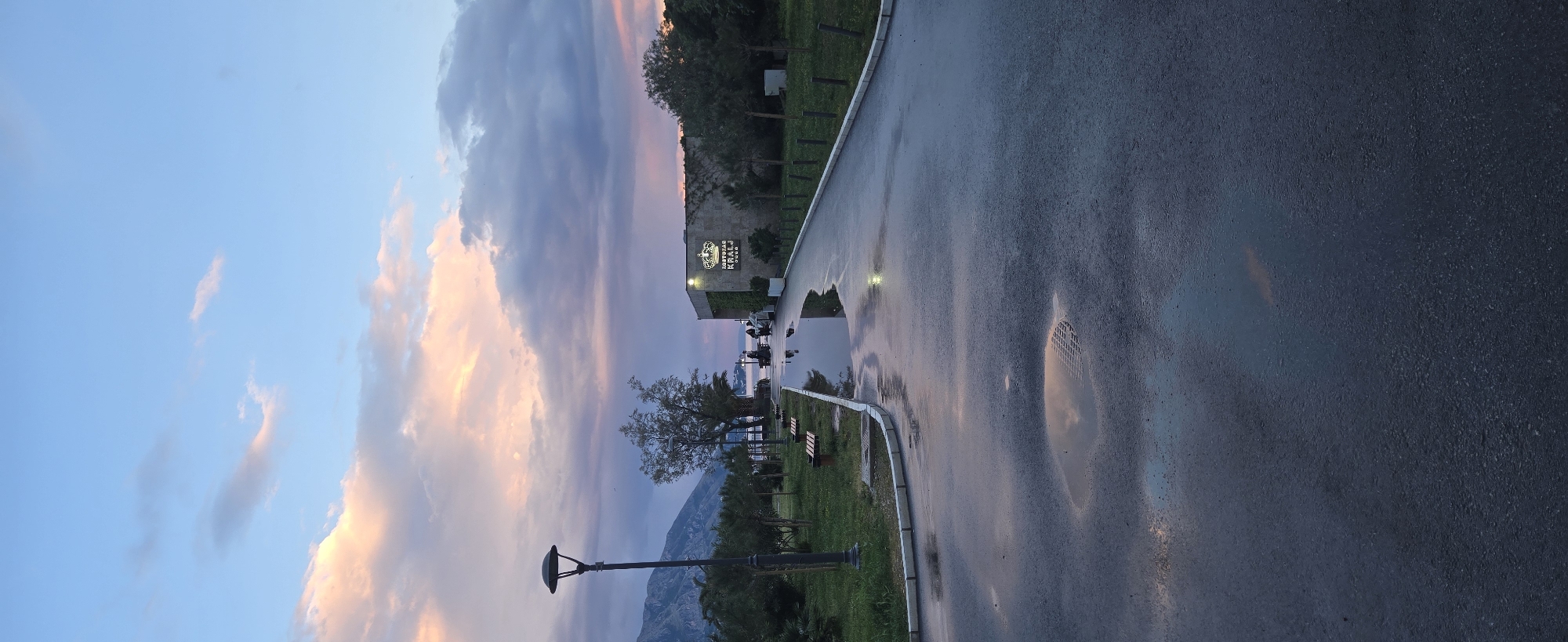 The image displays an outdoor evening scene in Budva, Montenegro, shortly after a rain shower, with the ground still wet and reflective. The sky is a prominent feature, transitioning from a clear blue at the top to a mix of soft pink, orange, and purple hues of scattered clouds, indicating either a sunset or dusk.
A wide, wet asphalt road or promenade occupies the right side of the frame, showing numerous reflections of the vibrant sky and artificial lights. On the left side, beyond a strip of green grass and trees, stands a building with a brightly illuminated sign. The sign features Cyrillic text, reading "РЕСТОРАН КРАЉ" (RESTAURANT KRALJ) with "CLUB" written below it, accompanied by a crown logo.
Various trees and lush green vegetation line the edge of the road and surround the building. In the foreground on the left, a modern streetlight with a dark, flat head is visible. In the background, dark, imposing mountains rise, silhouetted against the colorful sky. Several puddles on the wet surface create striking mirror-like reflections, including one distinct circular drain cover. A few indistinct figures can be seen near the restaurant building, possibly on a waterfront area beyond. The overall ambiance is tranquil and visually appealing, capturing the calm beauty of the landscape at twilight.