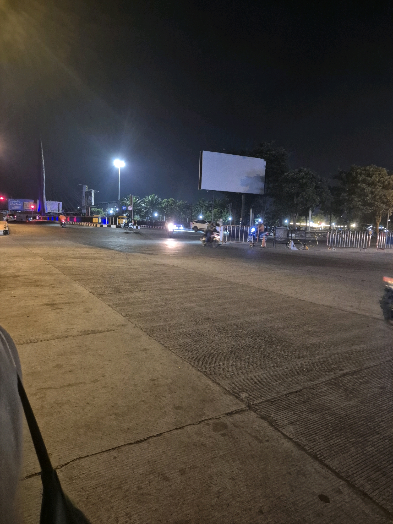 The image shows a nighttime street scene in Surakarta, Indonesia. The perspective is from ground level, looking across a wide, paved road. In the foreground, the concrete road surface is clearly visible, with some cracks and expansion joints. A dark shadow of a person's arm and bag is in the lower left corner.
The midground features a multi-lane intersection. Several motorcycles are in motion; one with an orange rider is moving away from the viewer towards the left, another is in the center of the frame, and a third is on the far right, approaching the viewer with its headlights illuminated. A white car is also visible on the right side of the road, with its headlights on.
In the background, traffic lights are visible, with red lights illuminated on the left. There are streetlights casting illumination on the road and surrounding areas. Trees are present behind the road, and some structures, possibly buildings or overpasses, can be seen. A large, blank billboard is prominently displayed in the upper midground. Metal barriers and traffic cones are set up along the side of the road. The overall impression is a busy urban intersection at night.