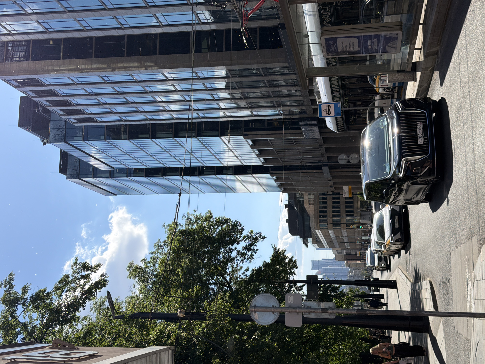 This image depicts a vibrant, sunny city street scene in Moscow, Russia, taken from a slightly elevated perspective. Bright daylight illuminates the area, casting strong reflections on glass surfaces and vehicles, while the sky is clear blue with scattered white clouds.
On the left, a large, modern skyscraper with a glass facade dominates the frame, its reflective windows mirroring parts of the sky and adjacent structures. Overhead, numerous wires crisscross against the sky and buildings.
The street itself is lined with vehicles. Most prominent in the foreground is a dark-colored luxury sedan, likely an Aurus Senat, identifiable by its distinctive grille and a Russian license plate displaying "C788CX 797". Further down the street, several white and yellow cars, characteristic of taxis or ride-share vehicles, are visible.
Along the right side of the street runs a sidewalk. A few pedestrians are present, including one individual with blond hair in the immediate foreground, possibly seated. Green trees with dense foliage are interspersed along the street, and in the distance, a skyline of additional tall buildings can be seen.
Street furniture includes various poles, a round street sign, and a bus stop shelter featuring a blue sign with a white bus icon. The glass panels of the bus stop shelter display text in Cyrillic script, with words like "Альфа" (Alfa) and phrases related to "how to pay" being partially legible, likely public information or advertisements. The scene conveys the atmosphere of a bustling, modern urban environment.