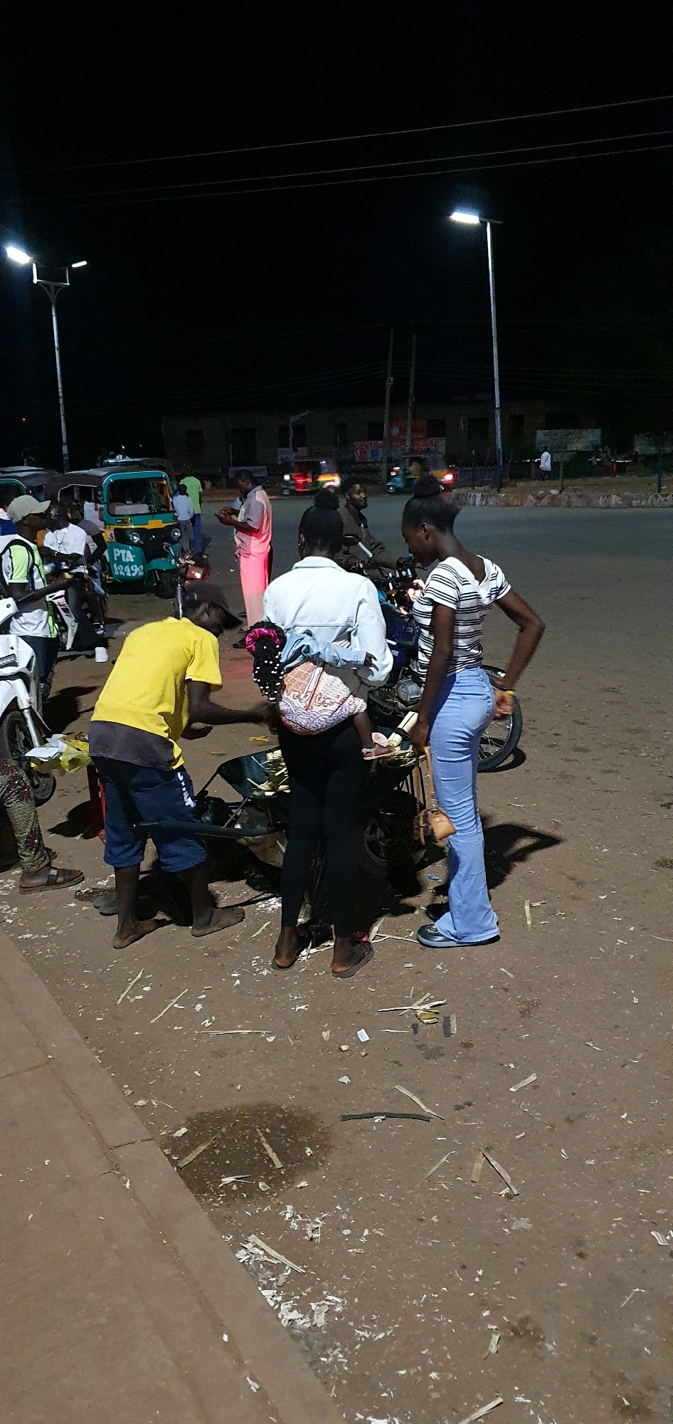 In Bukuru, Nigeria, at night, a group of people are gathered around a motorcycle on a paved road. A person in a yellow shirt stands to the left, wearing blue shorts and a yellow t-shirt. A woman in black leggings and a white denim jacket is in the center, holding a child wrapped in a patterned fabric on her back. A second woman on the right wears light blue flared pants and a striped t-shirt. A motorcycle is positioned between the individuals, with some debris scattered on the ground around it. In the background, streetlights illuminate the scene, and other vehicles, including a tuk-tuk, are visible. The overall environment suggests a public gathering or waiting area at night.