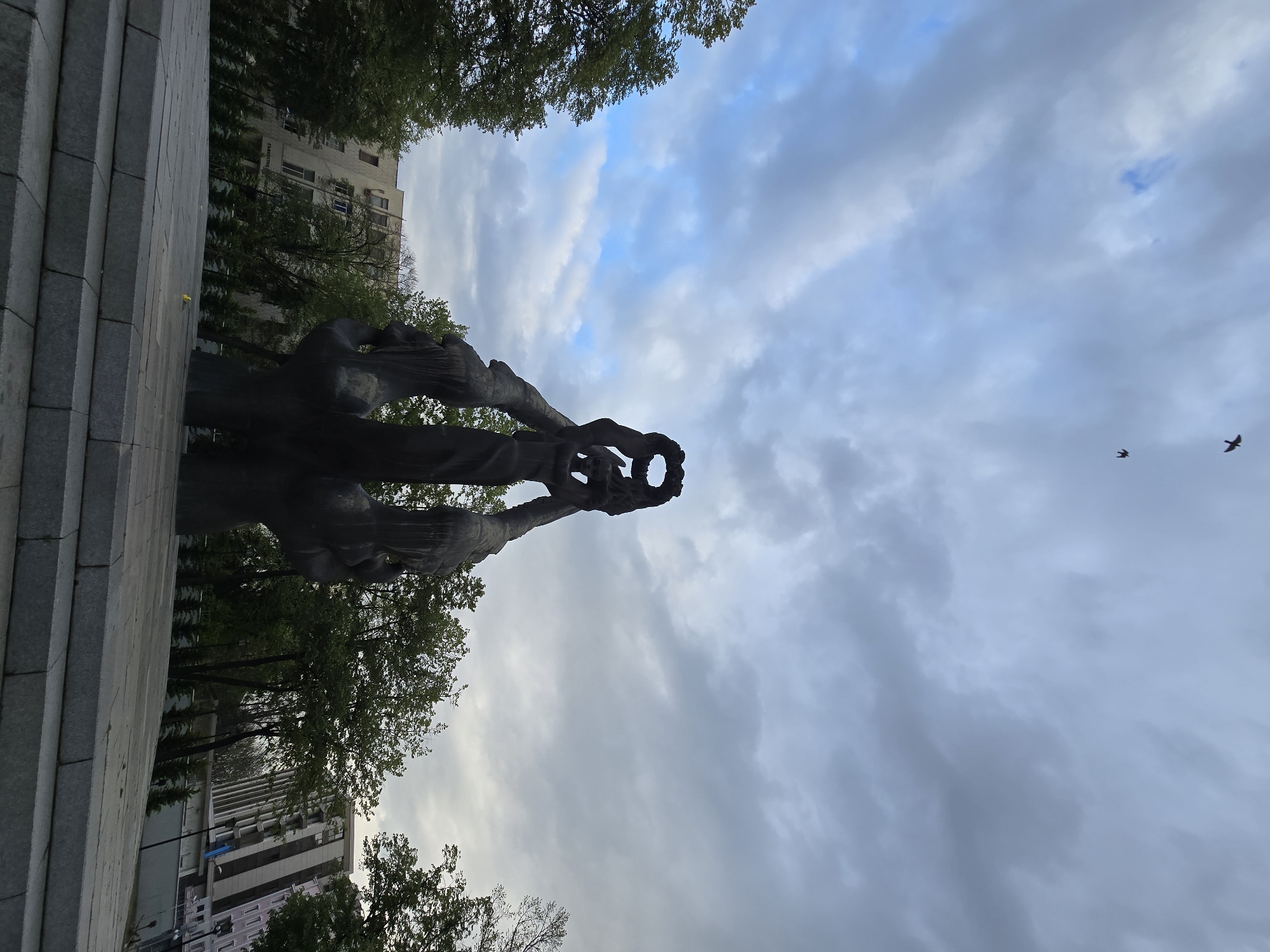 The image captures a powerful, dark-colored statue, likely made of bronze, set against an overcast sky in Plovdiv, Bulgaria. The statue is viewed from a low angle, emphasizing its upward reach. It depicts a figure or multiple intertwined forms holding a large, circular wreath or crown-like object high above their head. The silhouette of the statue conveys a sense of solemnity, triumph, or remembrance.
The sky above is filled with a mixture of grey and white clouds, with patches of brighter blue peeking through, indicating an overcast but not dark day. Two birds are visible in flight in the upper right quadrant of the sky.
In the background, behind the statue, are numerous green trees, suggesting a park or public square setting. Beyond the trees, parts of several multi-story buildings with light-colored facades are visible, typical of an urban environment. The base of the monument consists of large, grey, rectangular stone blocks. The lighting suggests it is daytime, possibly late afternoon or early morning, lending a slightly muted atmosphere to the scene. No discernible text is clearly legible on the statue or the surrounding buildings, though some indistinct characters might be present on a distant building.