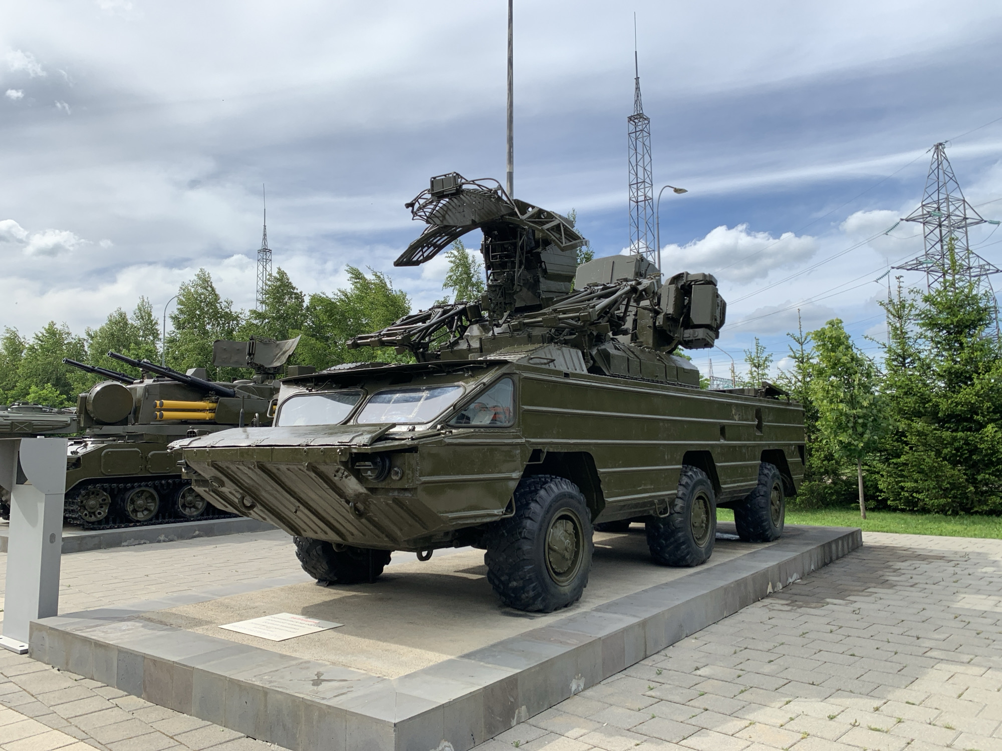 A green, wheeled military vehicle with a large radar dish mounted on top is parked on a paved area with concrete barriers. The vehicle has four large, knobby tires and a distinctive front design with a sloped, armored plate. Behind it, partially visible, is a tracked military vehicle with a turret and gun barrels. In the background, there are trees, power lines, and utility poles under a cloudy sky. The scene appears to be an outdoor exhibition or display of military hardware. There are no people visible in the image.
