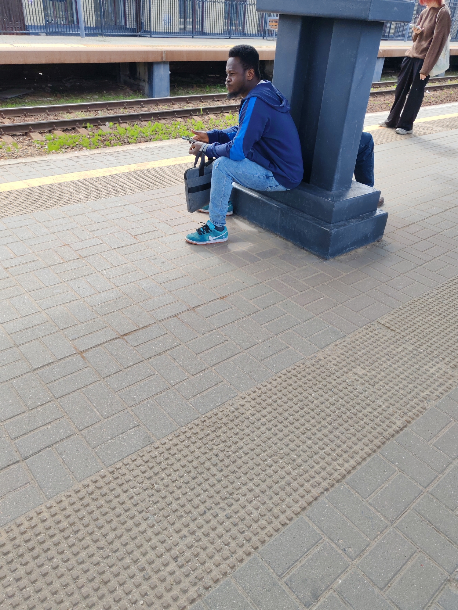 The image captures a scene on a train station platform in Moscow, Russia. The primary subject is a man with dark skin, short black hair, and a beard, who is seated on the platform, leaning against a dark grey, robust pillar. He is dressed in a blue hoodie with lighter blue details, light blue jeans, and distinctive turquoise/teal sneakers with white soles. He holds a dark grey tote bag in his lap and a smartphone in his right hand, looking towards the left side of the frame.
In the background, to the right and further down the platform, a second person is partially visible. This individual has light-colored hair and is wearing a brown sweater and dark pants, appearing to stand and also look at a smartphone.
The platform itself is paved with grey rectangular concrete blocks, arranged in a stretcher bond pattern. A tactile paving strip, featuring raised square studs, runs horizontally across the foreground, indicating the edge of the platform or a warning area. Beyond the platform where the man is seated, train tracks are visible, with patches of green vegetation growing between them. A raised concrete structure or a second platform, topped with a metal fence, is seen behind the tracks.
The scene takes place during bright daylight, suggesting a clear day. The activity depicted is primarily waiting at a train station. There is no visible text in the image.