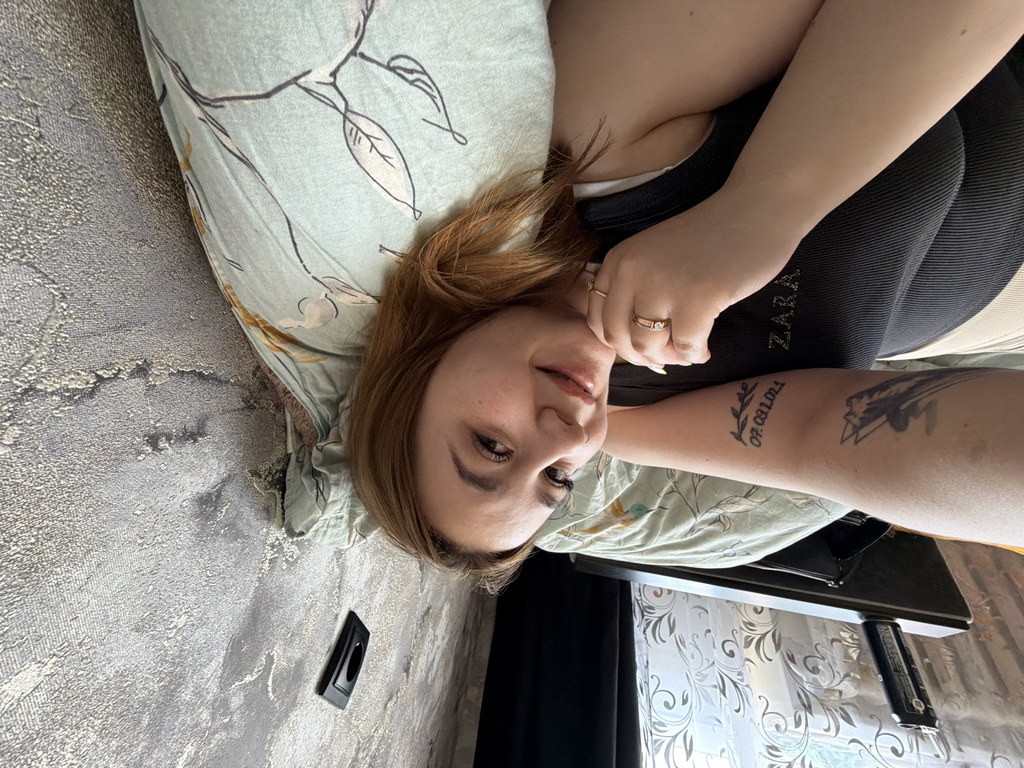 The image depicts a person lying down on a pillow with a floral pattern. The person has reddish-brown hair and is wearing a dark tank top with the word "ZARA" printed on it. They are wearing two rings on their left hand. Two tattoos are visible on their left forearm, one featuring a branch with leaves and a date "07.03.2021" and another of a mountain. The background includes a wall with a textured gray and gold pattern and a black electrical outlet. Next to the bed is a small table in front of a window with a decorative curtain. The location is Chaikovsky, Russia.