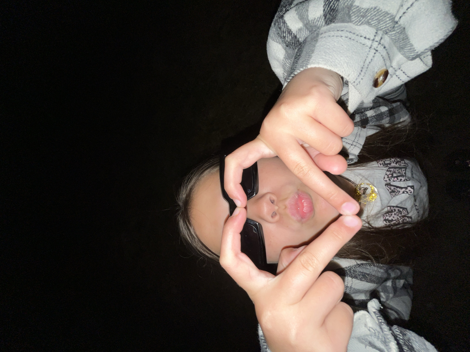 A person is posing for a photograph. They are wearing sunglasses and making a heart shape with their hands around their face. They are wearing a grey and white plaid shirt. The background is dark. The location is Novopokrovskaya, Russia.