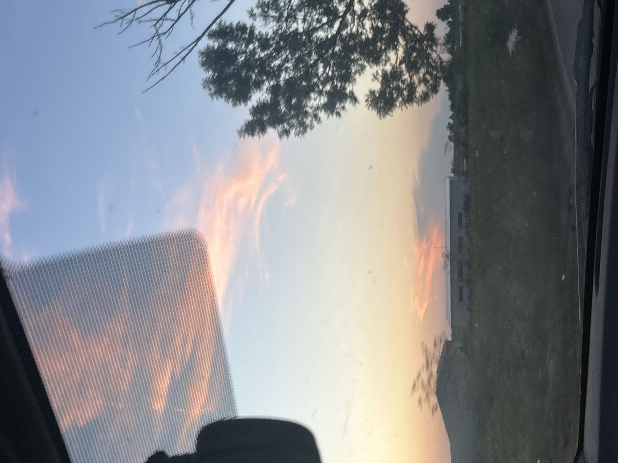 The image captures a sunset scene viewed through a car windshield. The sky is a gradient of blue, transitioning to soft pink and orange hues where the sun is setting. Wispy clouds are scattered across the sky, catching the warm light. In the foreground, the textured pattern of the windshield is visible, along with a portion of the car's interior.
The landscape beyond the windshield features rolling hills covered in green vegetation. In the middle ground, there are several buildings, appearing to be industrial or commercial structures, one with a long, low profile and another with a sloped roof. Silhouetted against the sky are the branches of a large tree to the right of the frame, and the indistinct shape of another tree or structure to the left. The overall impression is one of a rural or semi-rural setting at dusk. The image is slightly blurry, suggesting it was taken while the vehicle was in motion.