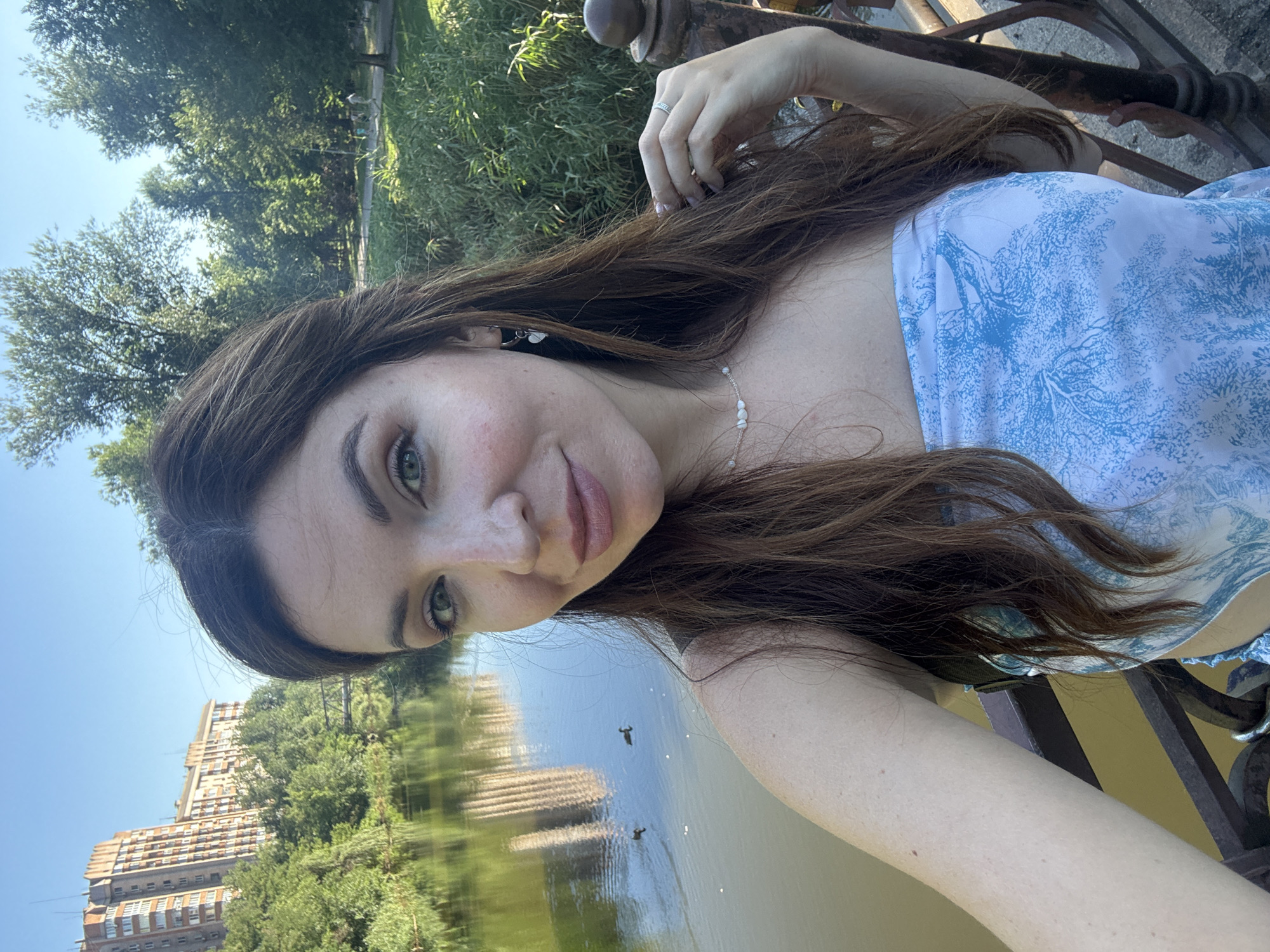 A woman is taking a selfie outdoors in Kryvyi Rih, Ukraine. She is standing on a bridge overlooking a body of water, with trees and buildings in the background. The water reflects the sky and buildings. There are two ducks swimming in the water. The woman has long brown hair and is wearing a strapless blue and white patterned top. She is also wearing a necklace and earrings. Her right hand is raised, touching her hair, and she is wearing a ring on her ring finger. The sun is shining, casting shadows and highlighting the greenery.