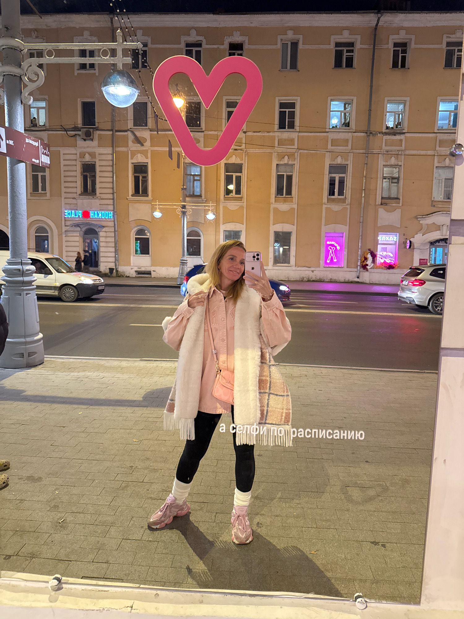 A woman is taking a selfie in a shop window at night in Tver, Russia. She is wearing a pink collared shirt, a fluffy white scarf, black leggings, white leg warmers, and pink sneakers. She is holding her phone in her right hand and smiling at the camera. In the reflection of the window, a pink heart decoration is visible, along with a street with cars and buildings. One building has signs that read "HOOKAH PLACE" and "NAKAZ". The street appears to be paved with cobblestones.