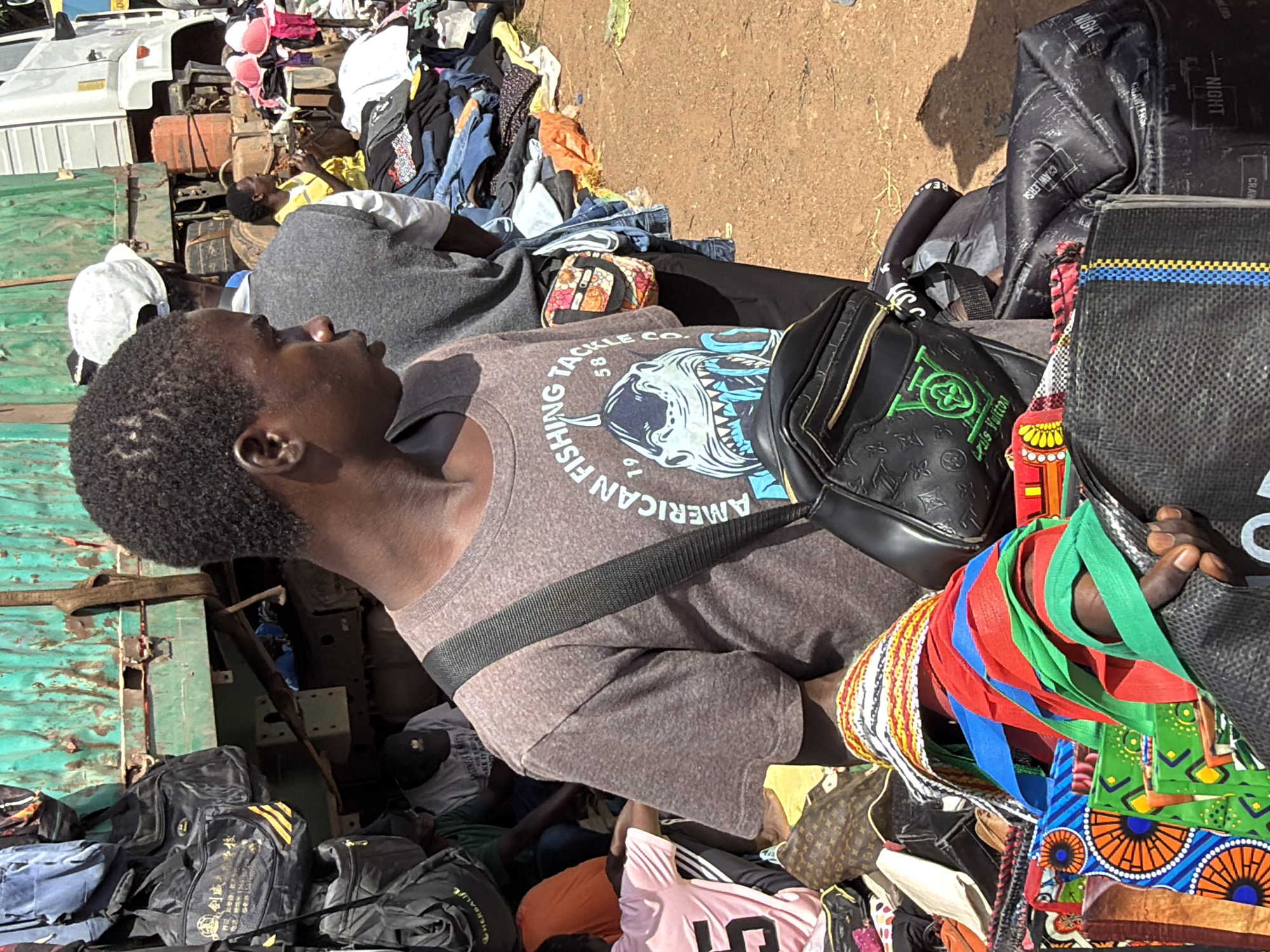 A young Black male with short, dark, curly hair and a brown t-shirt with a shark graphic stands in the foreground, facing right. He wears a dark crossbody bag with a green logo and the words "Louis Vuitton" on it. Behind him, a crowd of people is visible near a green truck. To the right of the truck is a pile of clothing and bras. The ground is covered in dirt. The scene appears to be a market in Gura Top, Nigeria.