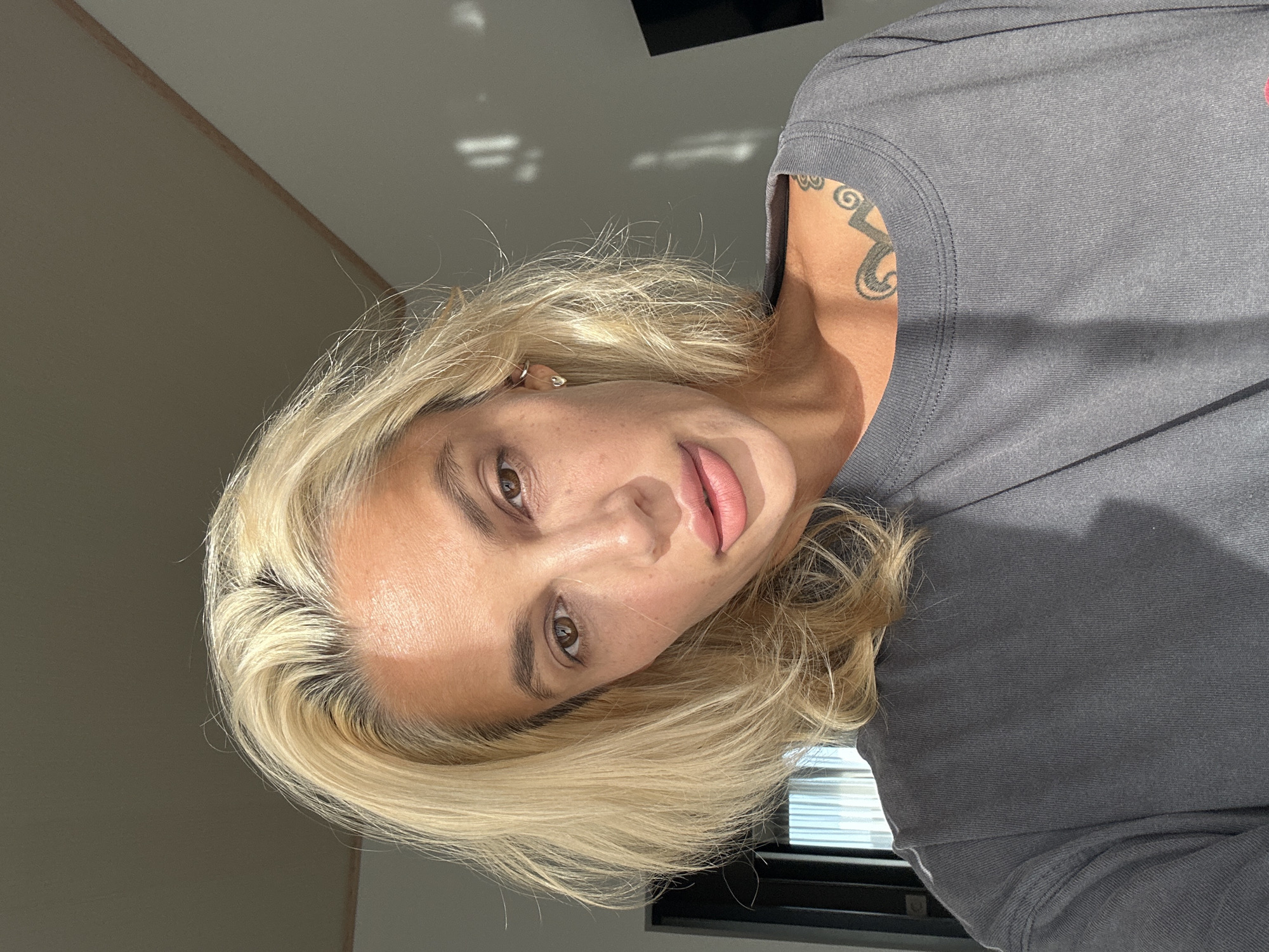 A mid-shot, overhead portrait of a woman with blonde hair and light skin is captured in natural lighting. She is wearing a dark gray t-shirt. The background shows a room with walls and a window with blinds. Light is casting shadows on the wall. She has a tattoo on her upper chest. The scene appears to be indoors, possibly in a residential or hotel setting.