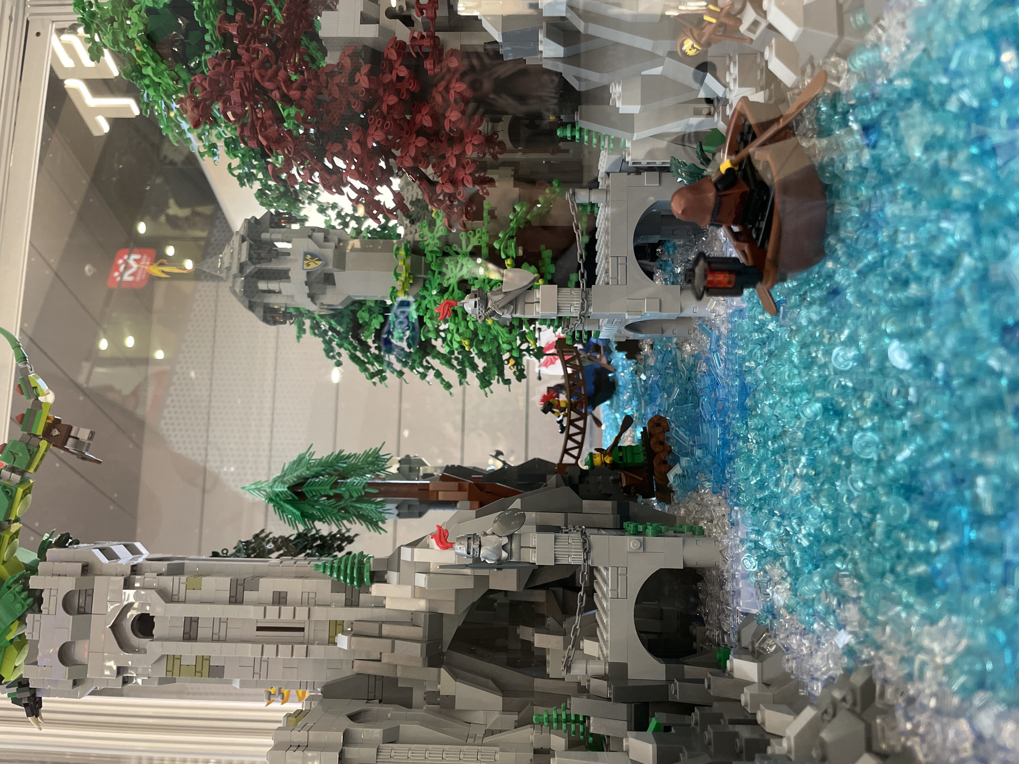 The image shows a detailed LEGO diorama depicting a medieval-fantasy scene set in a rocky, wooded canyon with a flowing river. The diorama includes two prominent stone castles with flags, one on the left and another in the background. Several small boats with figures are navigating the bright blue, translucent LEGO river. One boat in the foreground has a single figure rowing and a lantern attached to the back. The left castle is large and gray, built into a cliff face with an arched entrance. A knight figure in armor stands guard on a ledge of this castle. The background castle is smaller and gray with a flag on top. Lush green and red LEGO trees are scattered throughout the scene, adding to the natural environment. The diorama is displayed behind glass, with reflections of indoor lighting and some text visible on the glass, including a partially visible "TE" logo. There is no information in the image to suggest its location in the city of Sofia, Bulgaria.